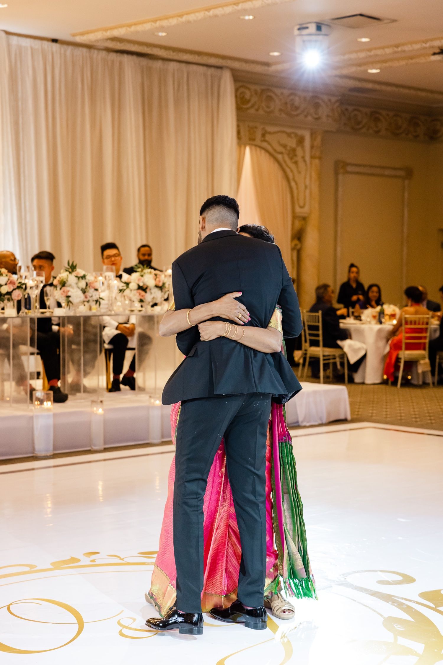 Couple embraces during romantic slow dance at their wedding reception with elegant table settings visible.