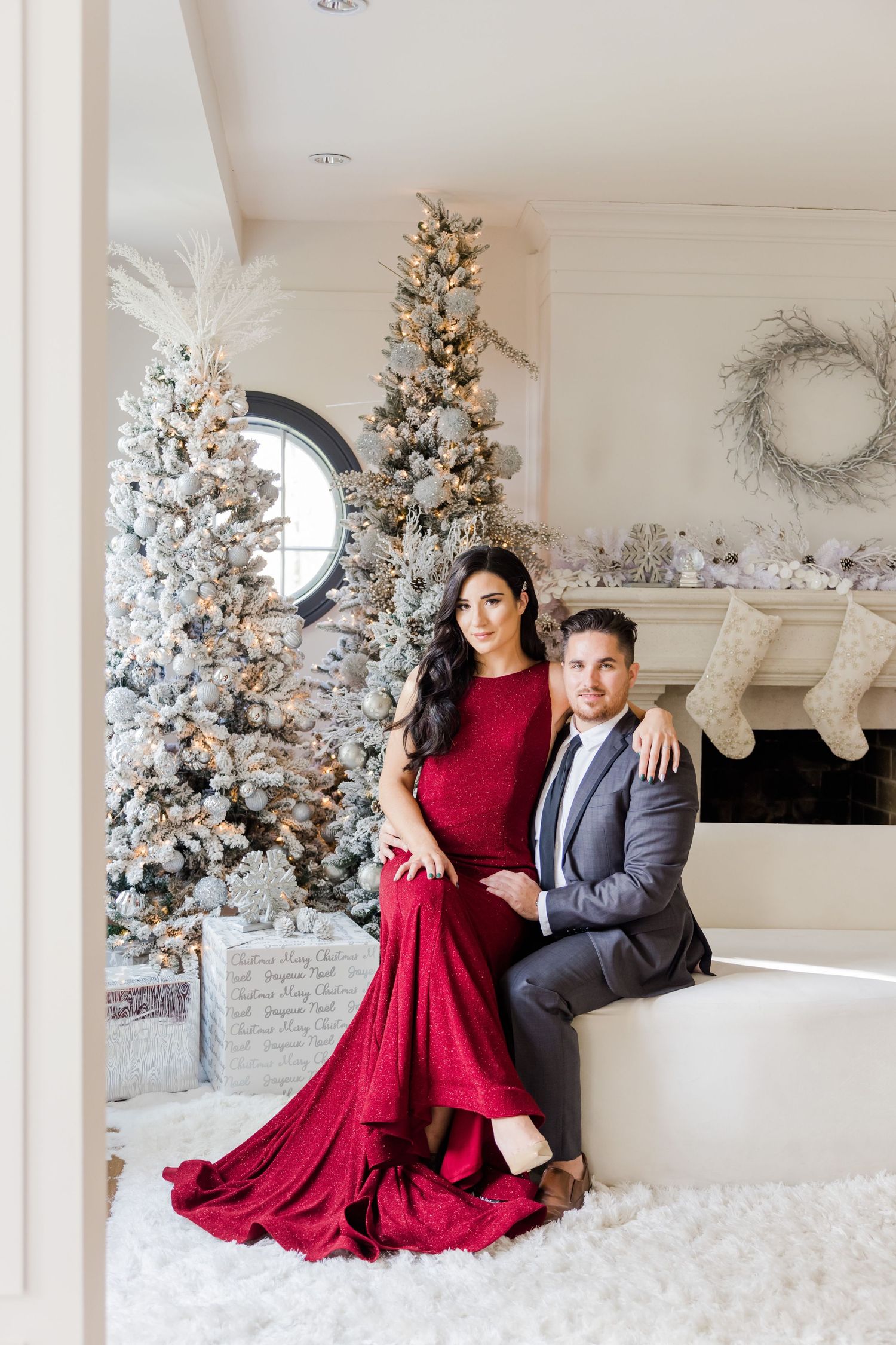 Formal couple's portrait featuring flowing red gown against snow-flocked Christmas trees and white decor.
