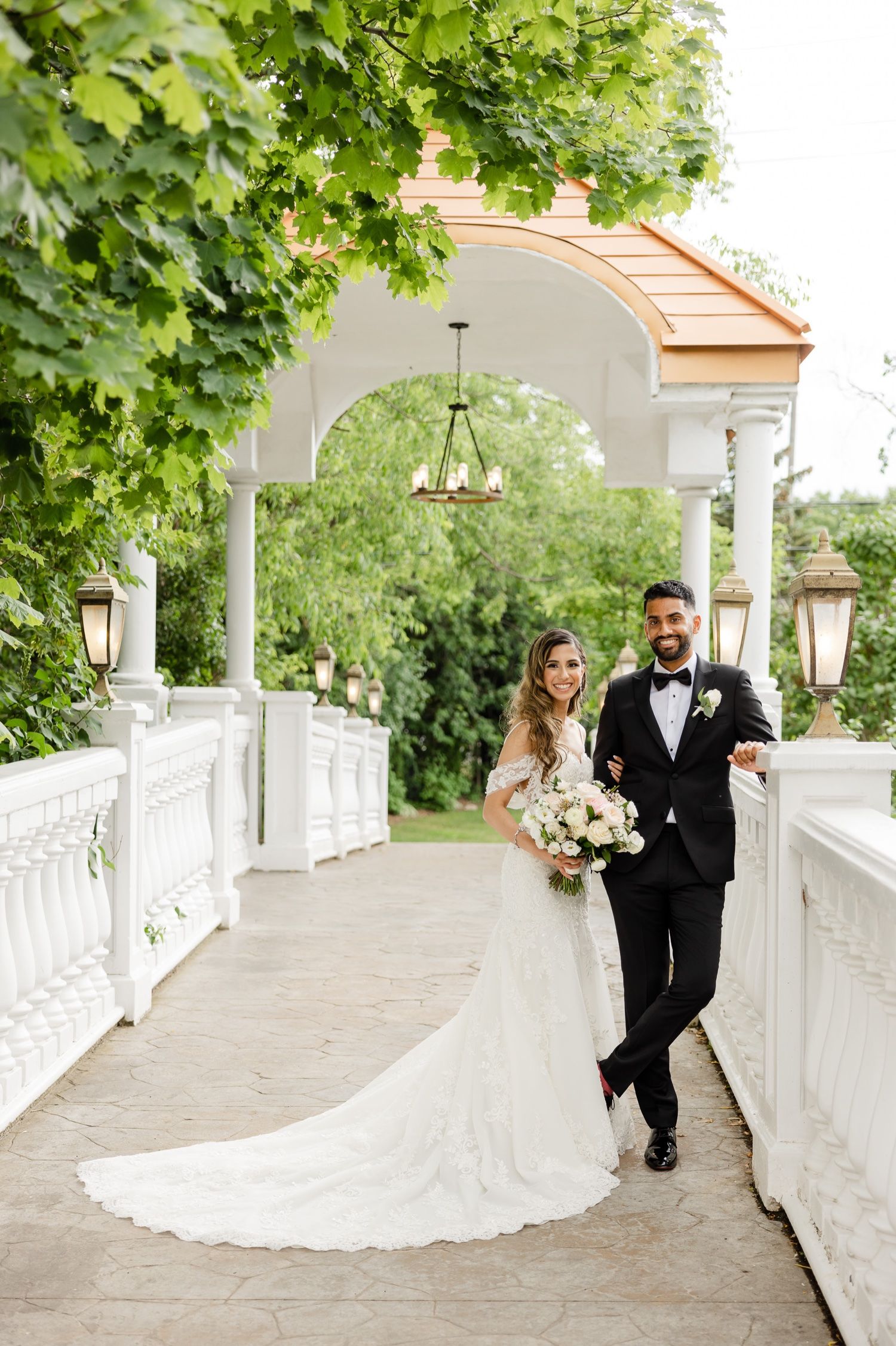 Wedding couple strolls along elegant white columned pathway under arched ceiling and greenery.