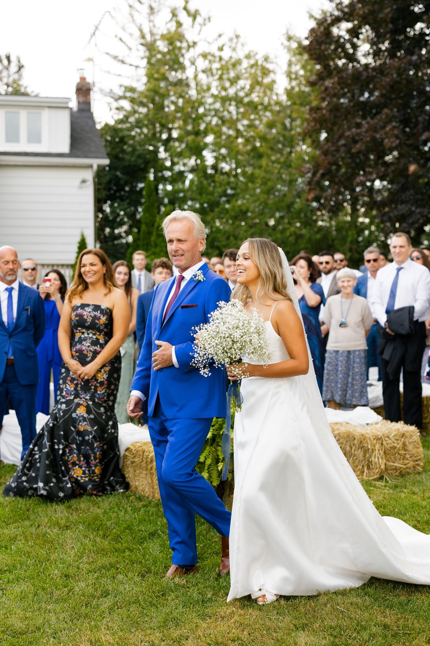 A formal wedding procession walks across a grassy lawn with guests looking on and a white house in the background.