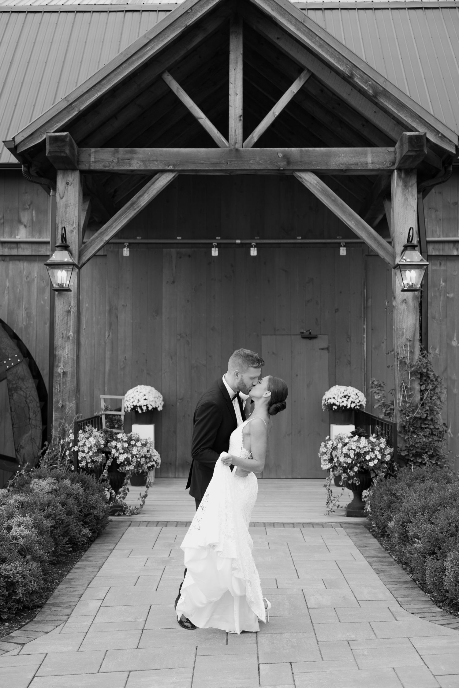Black and white romantic portrait at barn entrance framed by blooming flowers and lanterns.