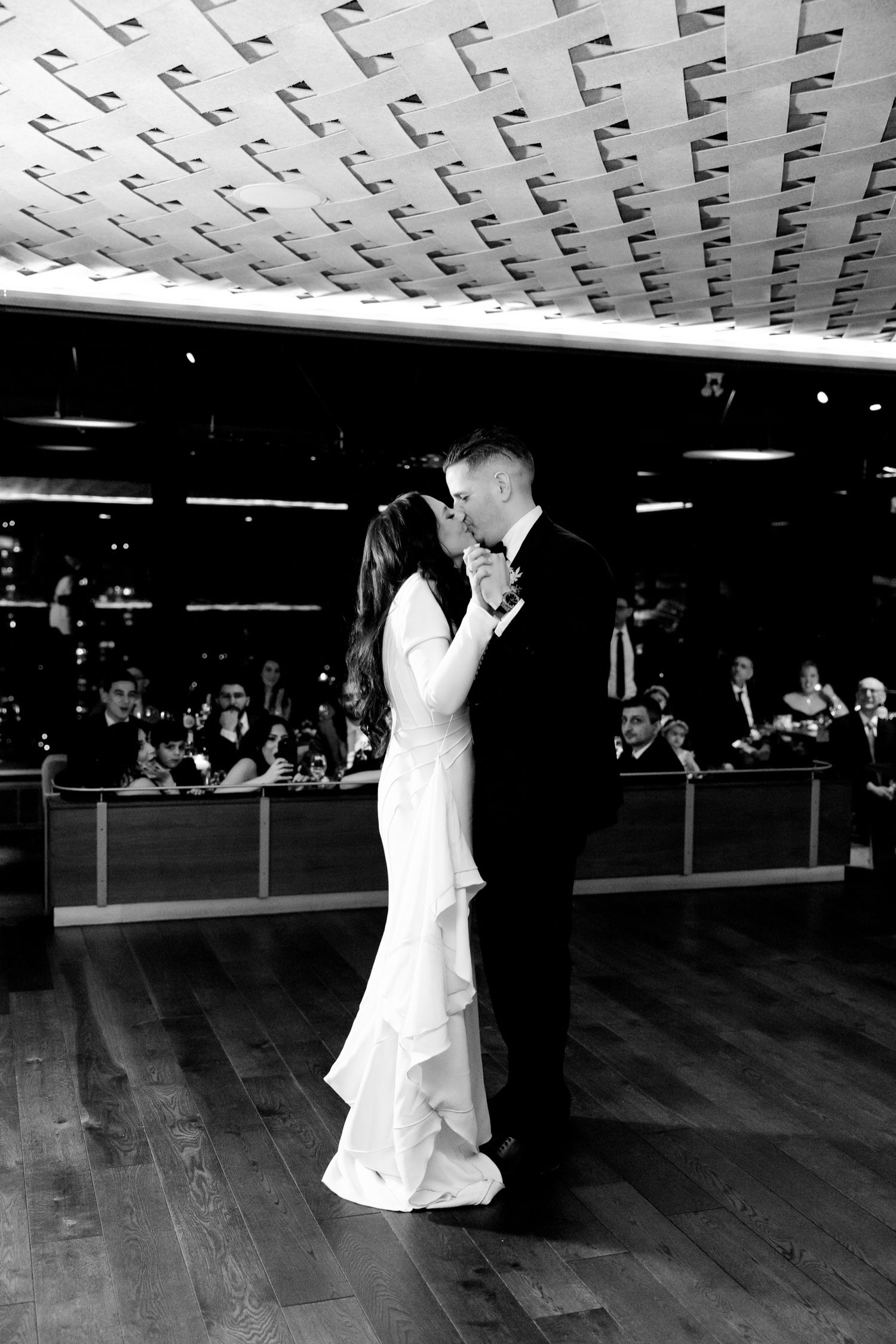 Black and white photo of dancing couple beneath geometric patterned ceiling with onlookers.