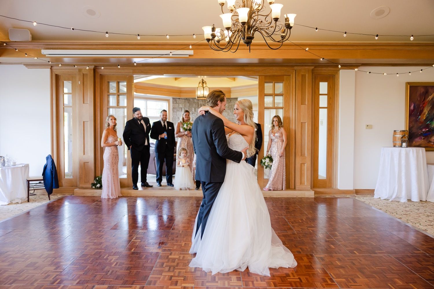 Couple sharing their first dance in an elegant ballroom with wooden accents and chandeliers.
