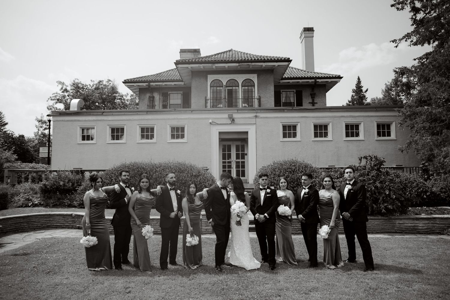 Wedding party poses in front of elegant white mansion in classic black and white photograph.
