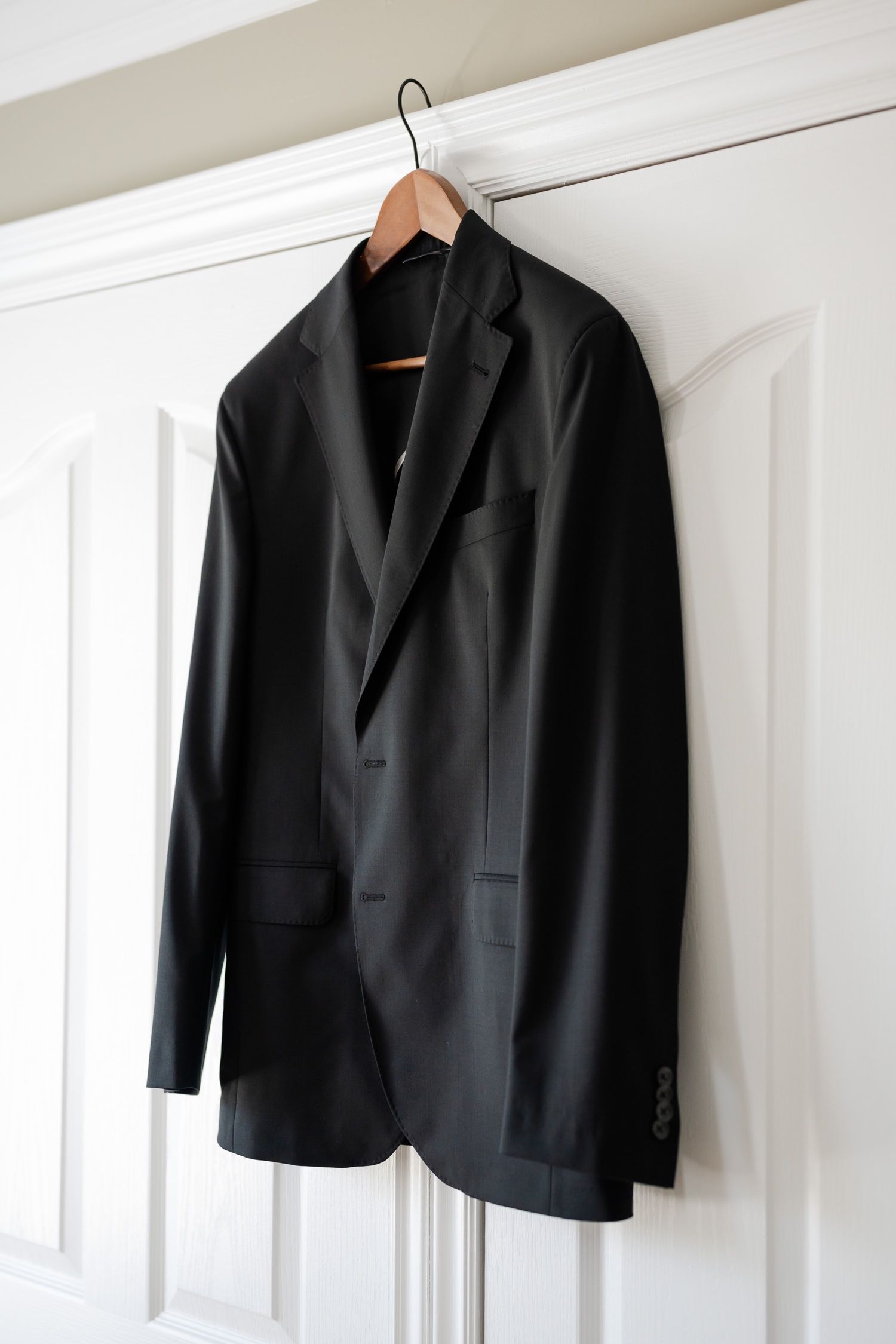 A black suit jacket hanging on wooden hanger against white door and wall.