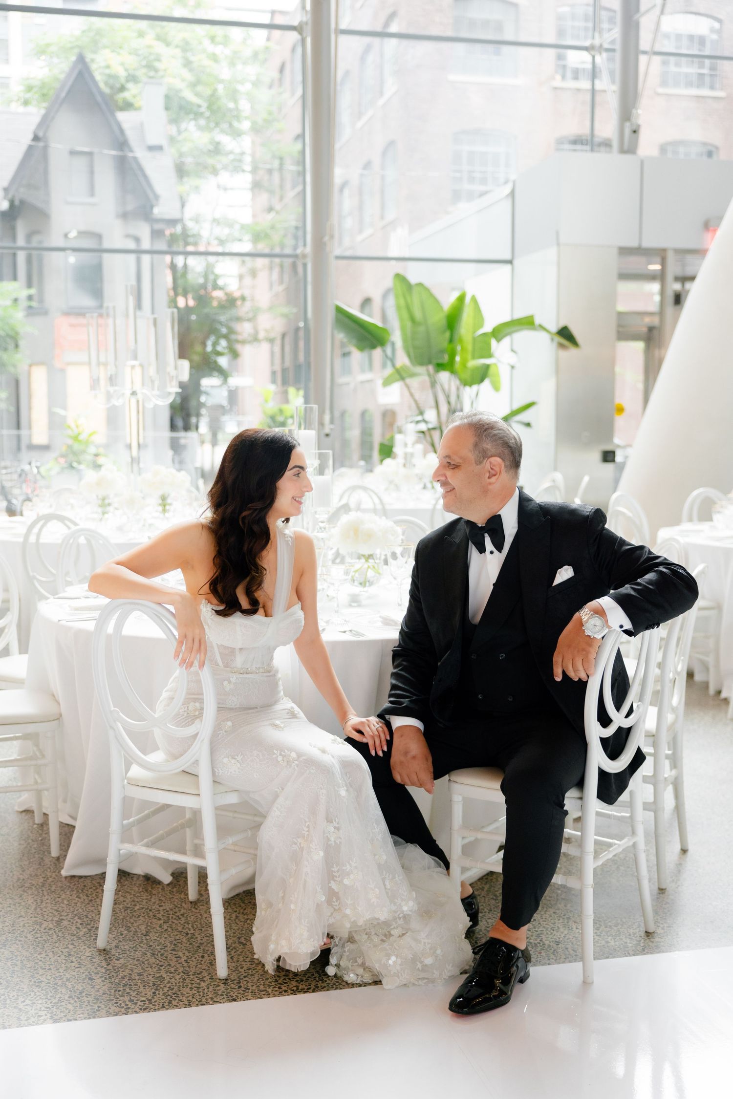 Wedding couple relaxing in white chairs at bright modern reception venue with large windows.