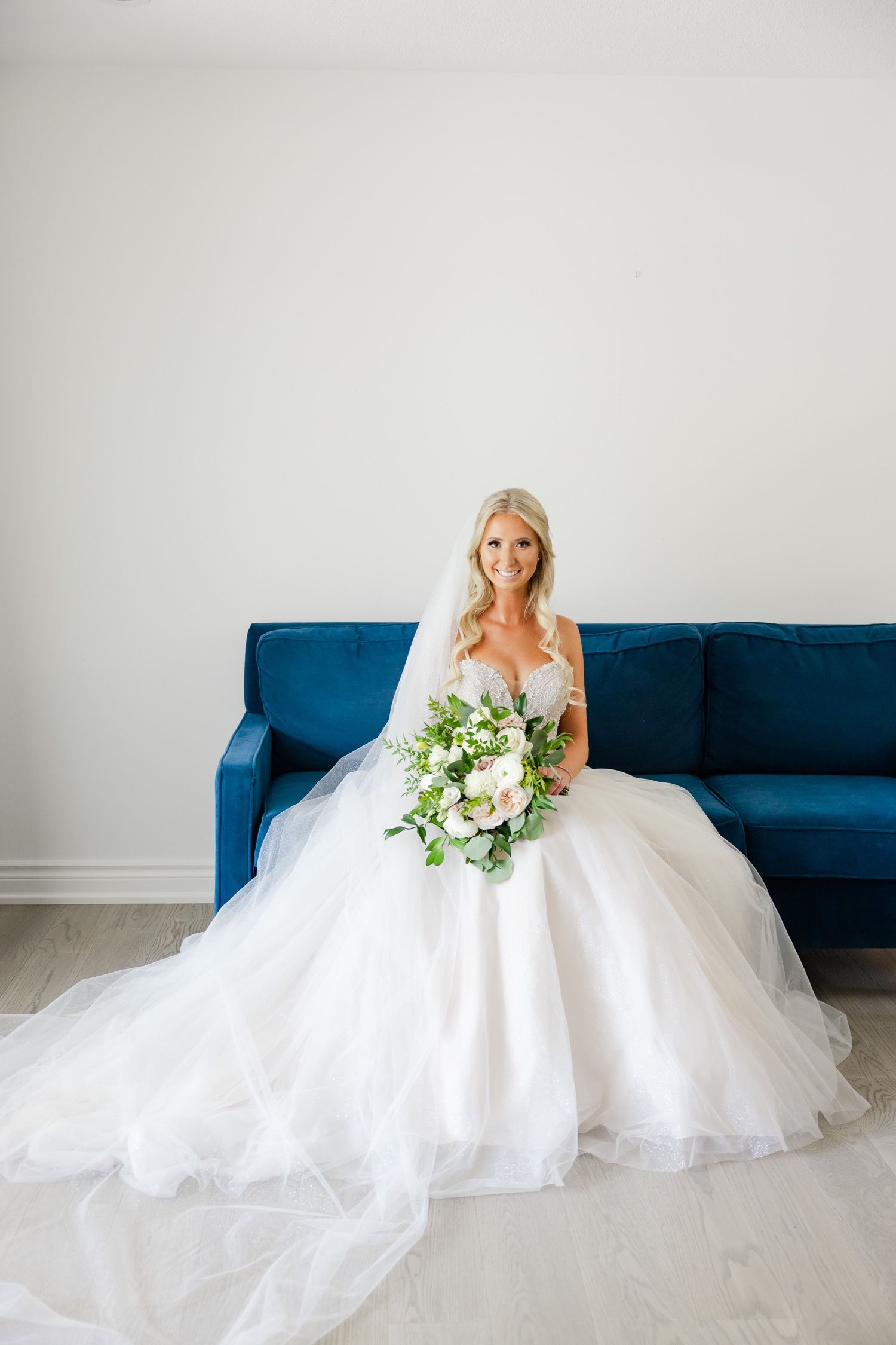 Elegant bride in flowing white gown poses with fresh bouquet on royal blue velvet sofa.