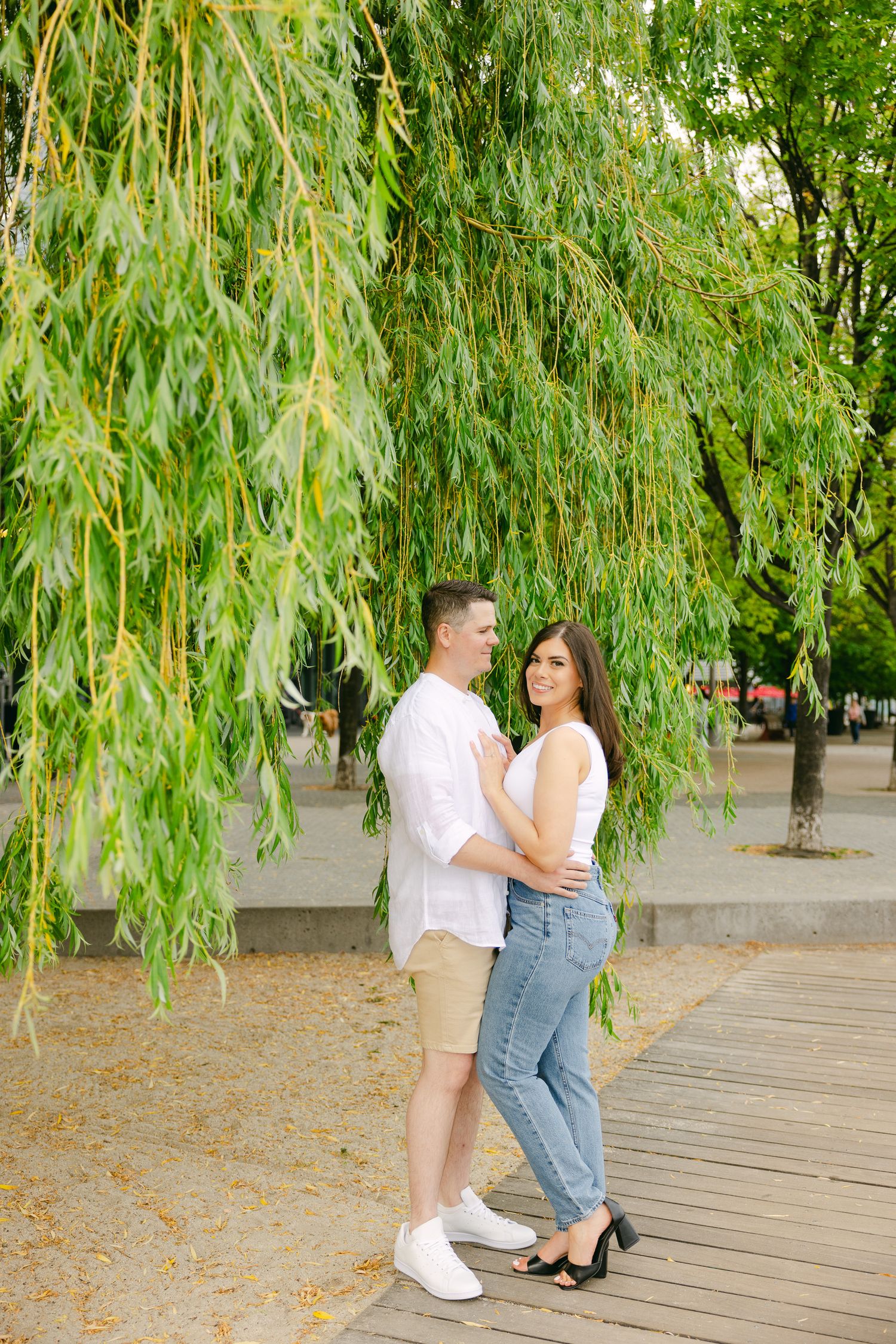 Stylish couple embracing on boardwalk beneath drooping willow branches.