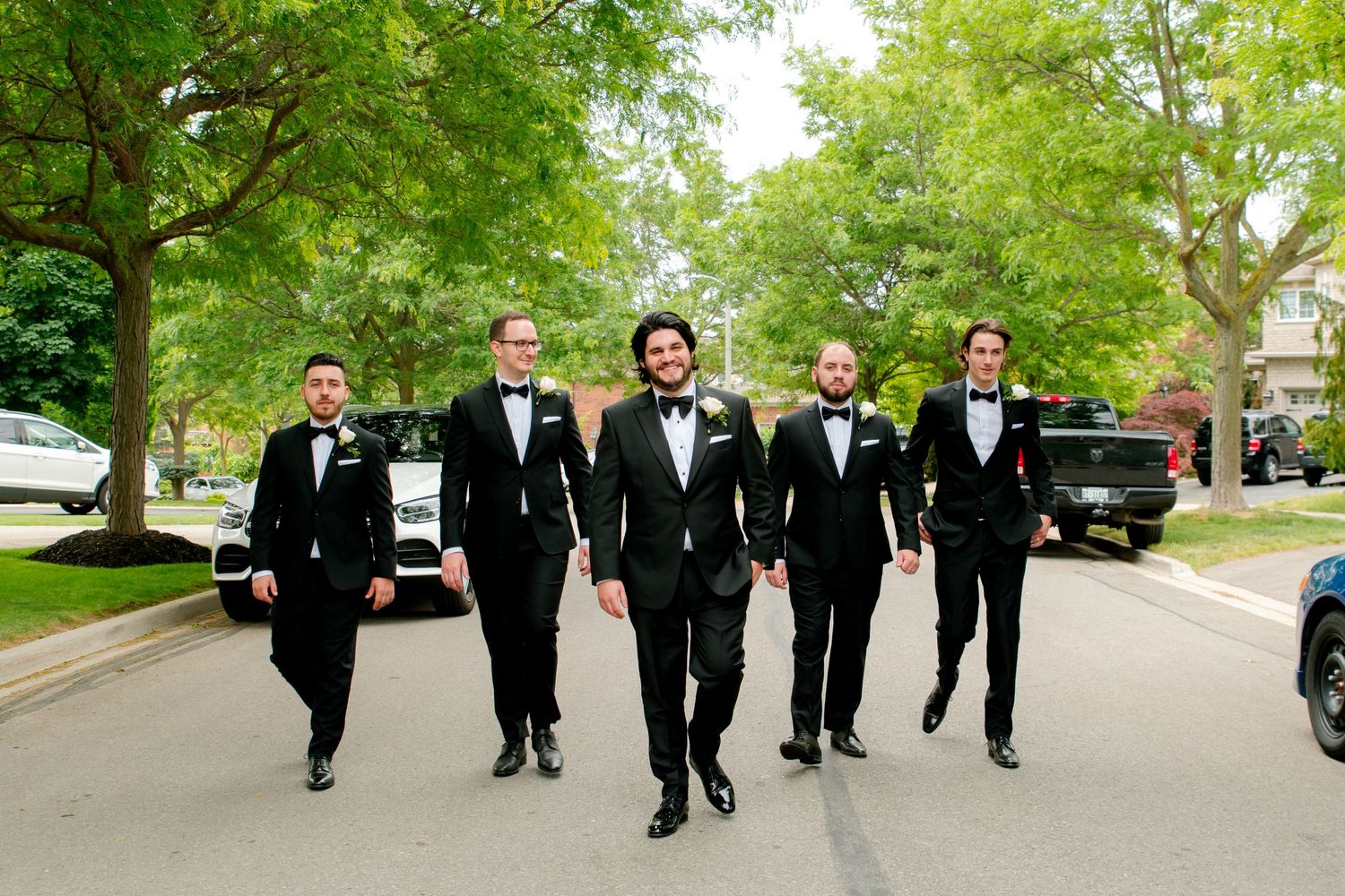 Five groomsmen walk together down a tree-lined street wearing black tuxedos before a wedding ceremony.