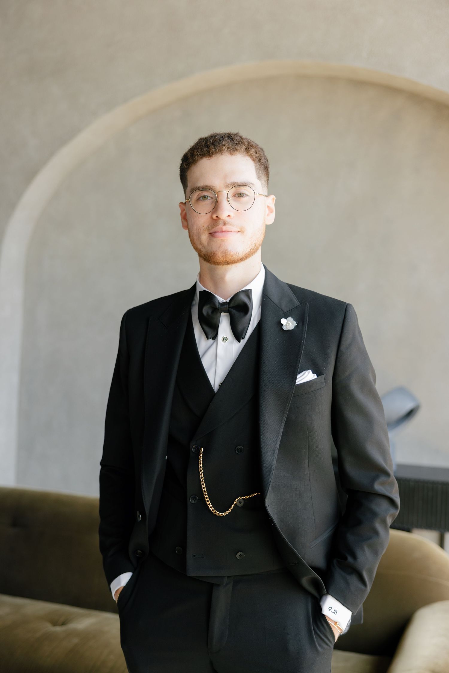 A formal portrait of a groom in a classic black tuxedo with bow tie and glasses against a neutral backdrop.