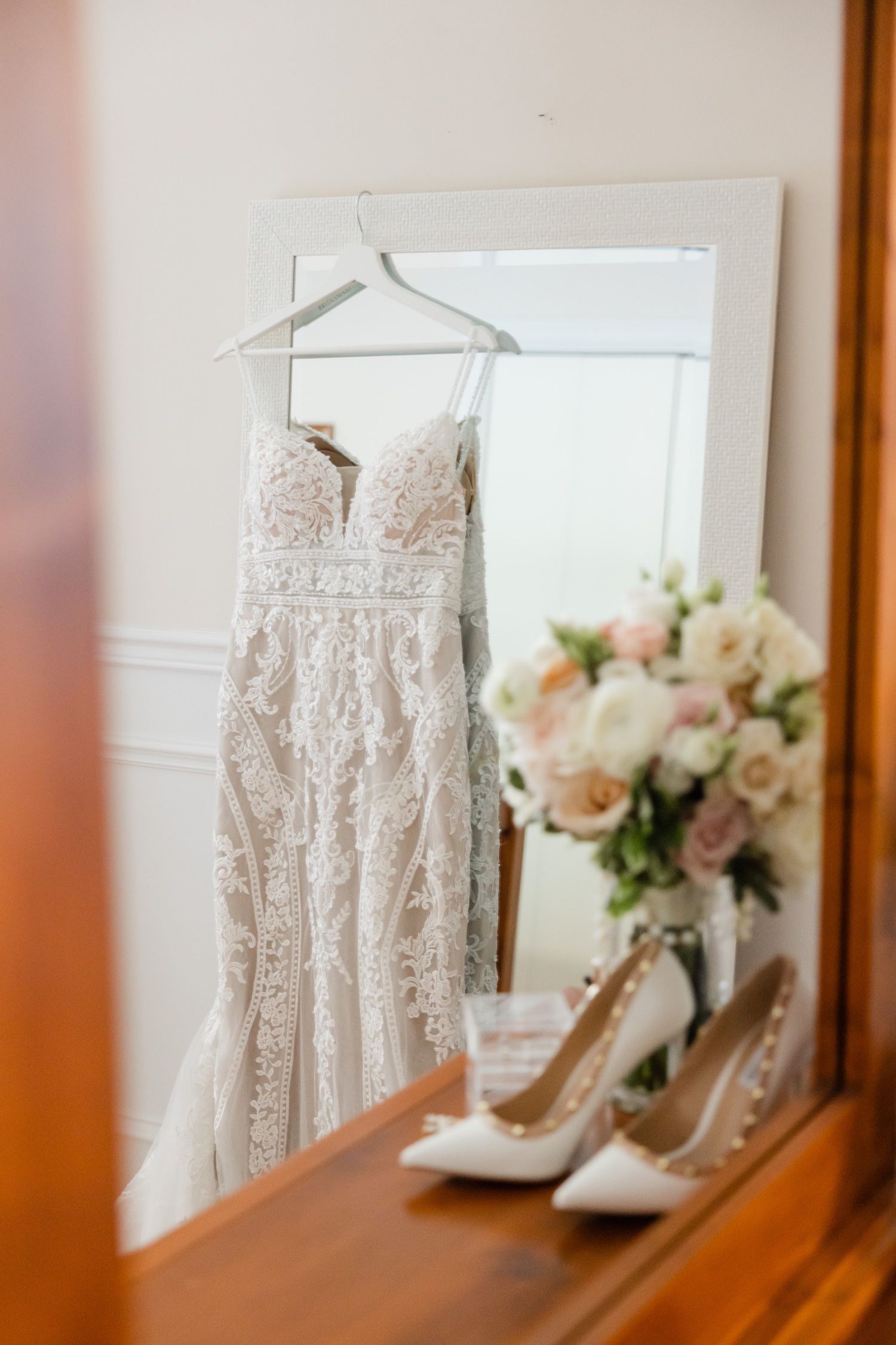 Elegant lace wedding dress hanging on white hanger with bridal shoes displayed below.