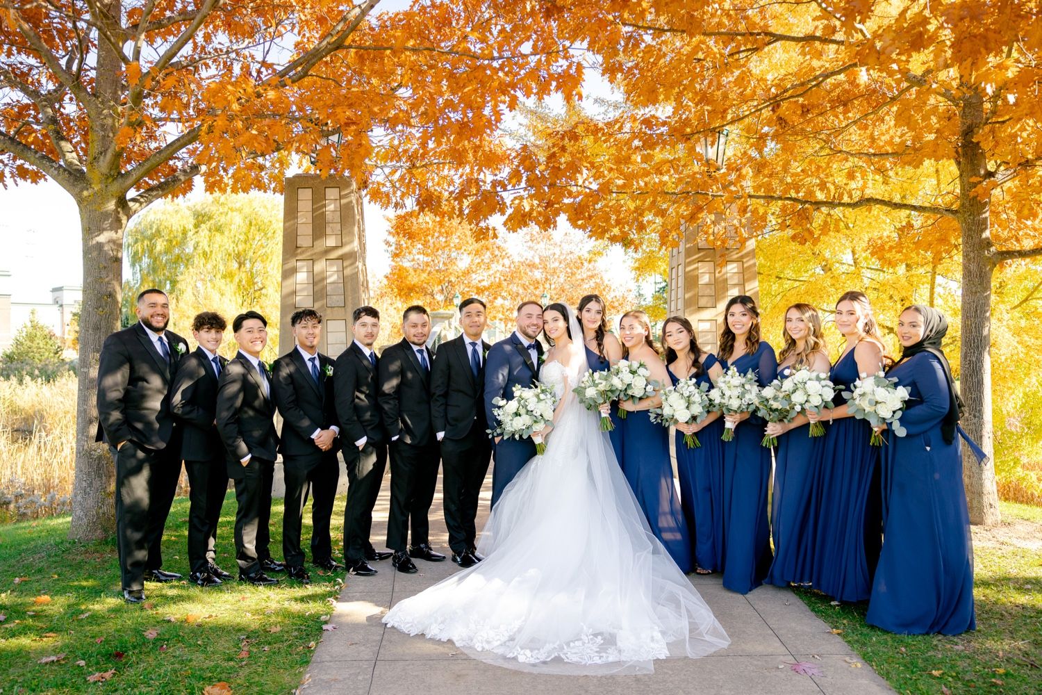 Wedding party in navy suits and royal blue dresses poses beneath vibrant autumn trees.