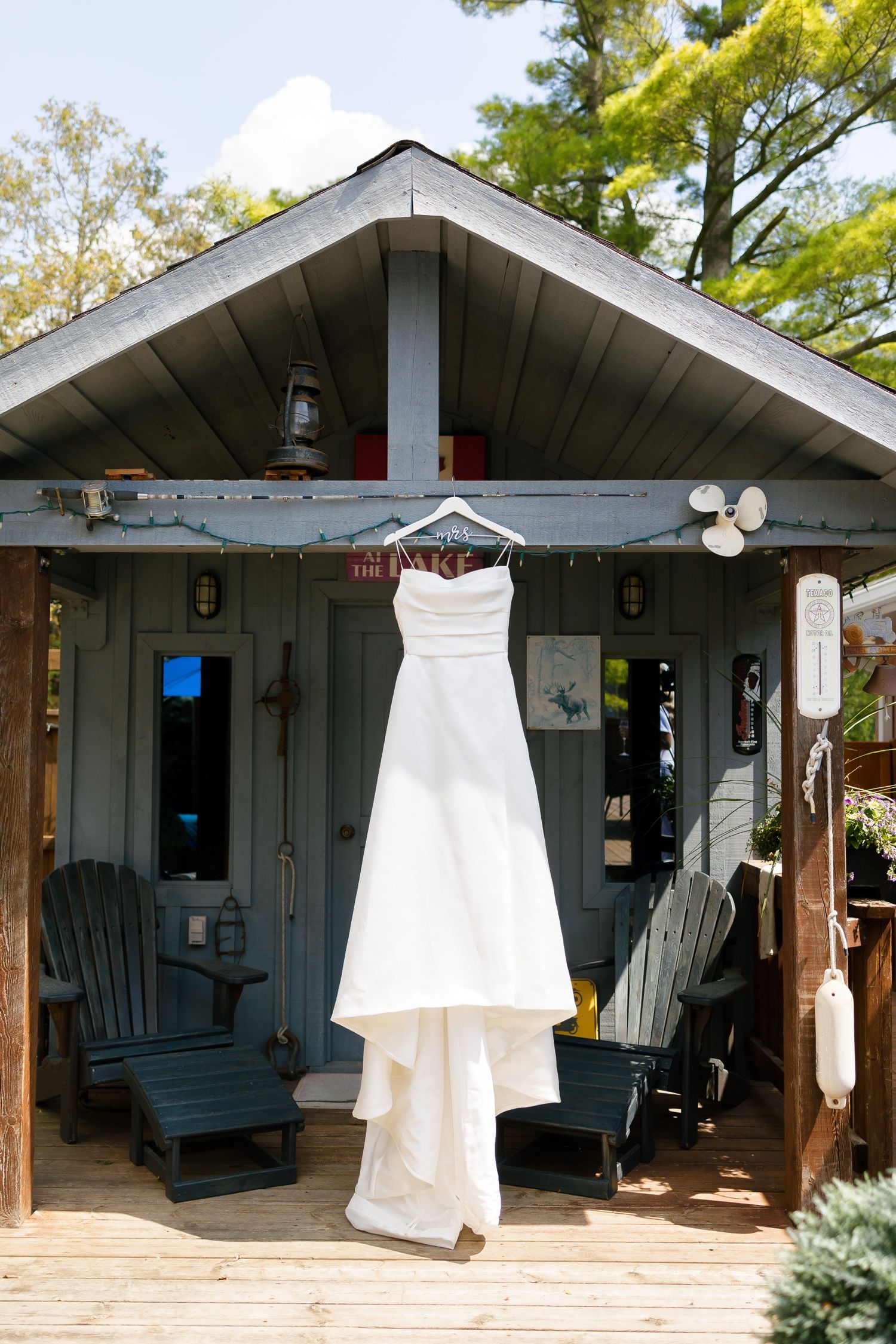 White wedding dress hanging on display outside a rustic cabin with blue rocking chairs.