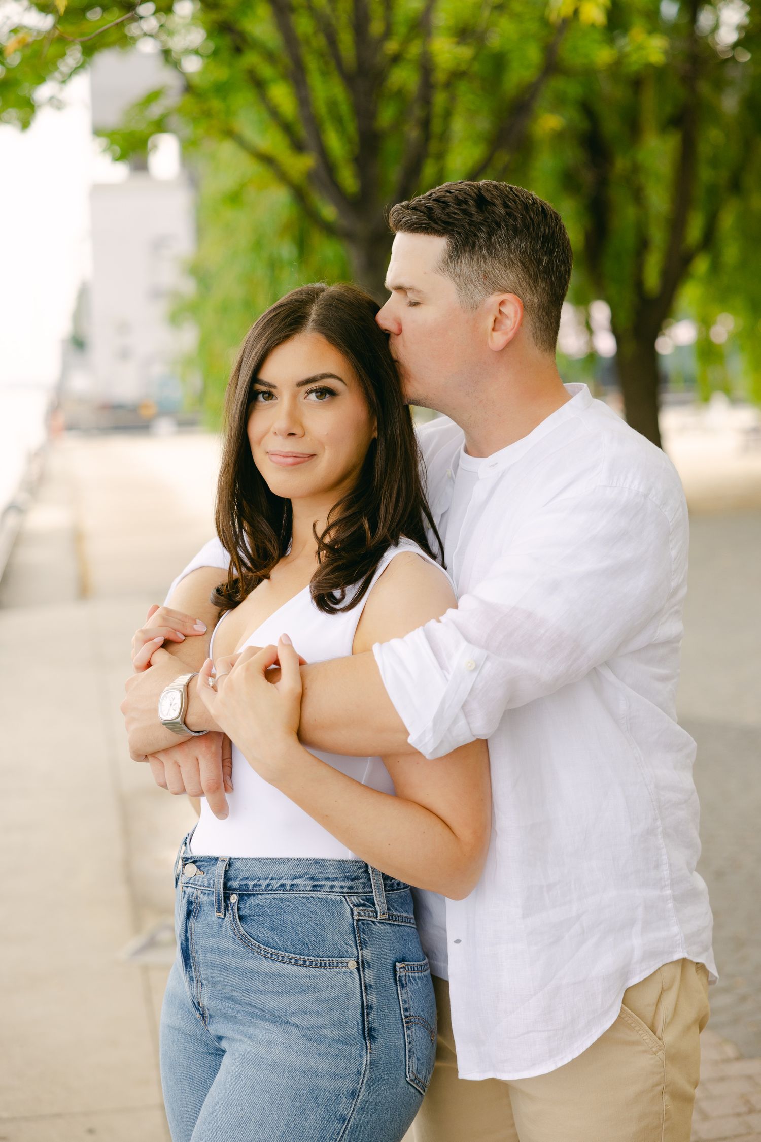 A tender moment captured between a couple under green trees in casual summer attire.