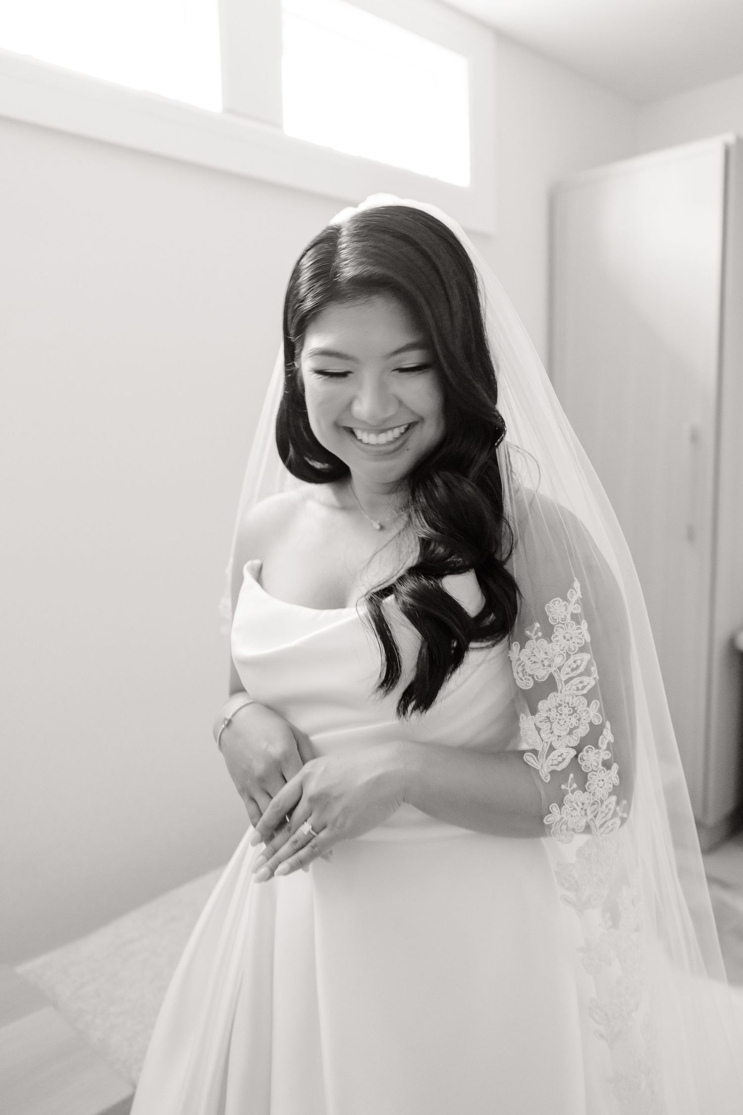 A radiant bride in a white gown and veil smiles joyfully while looking down during her wedding preparations.