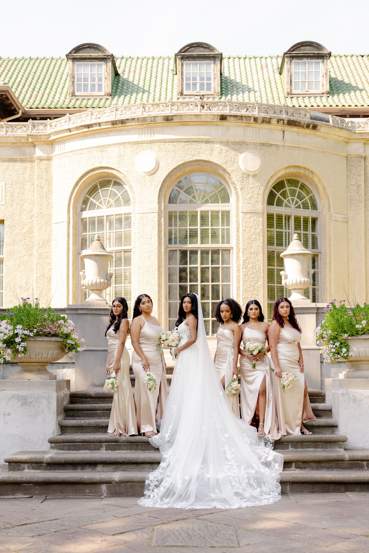 Bridesmaids in champagne gowns gather on stone steps with the bride wearing a lace wedding dress and long train.