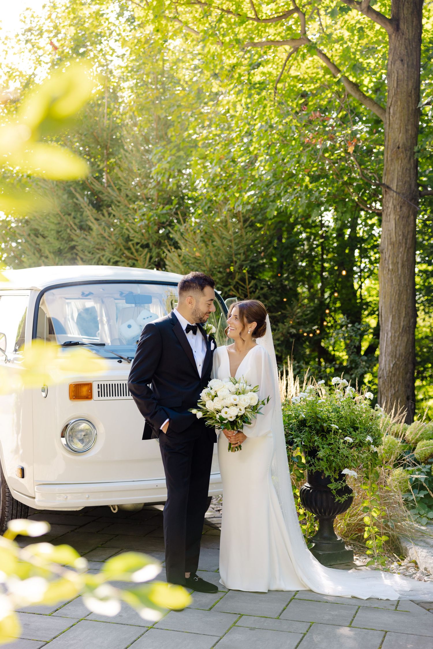 Elegant wedding couple poses by vintage white VW van amid sunlit garden setting.