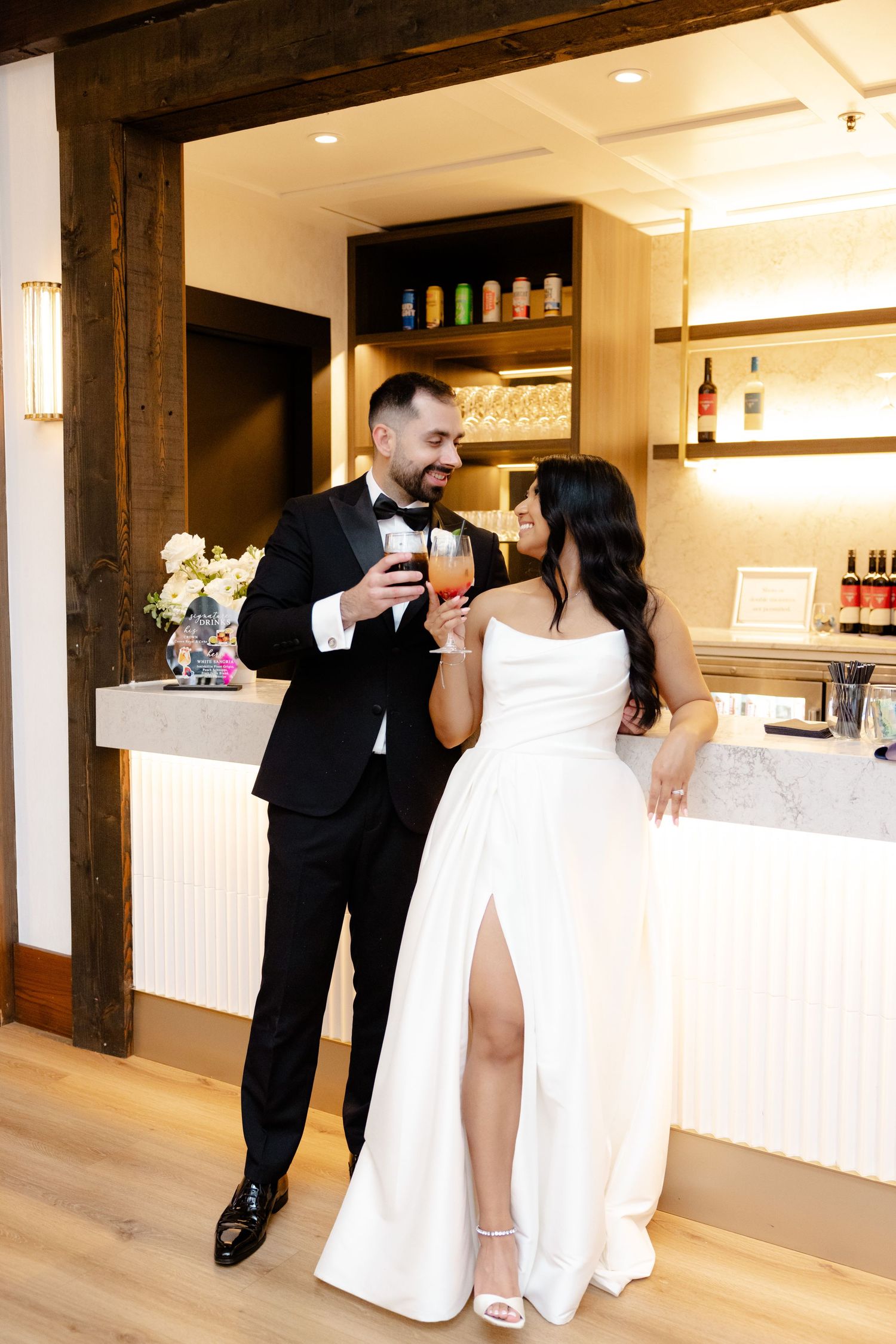 Newlyweds share a toast at a stylish bar area decorated with wooden beams and glass shelves.