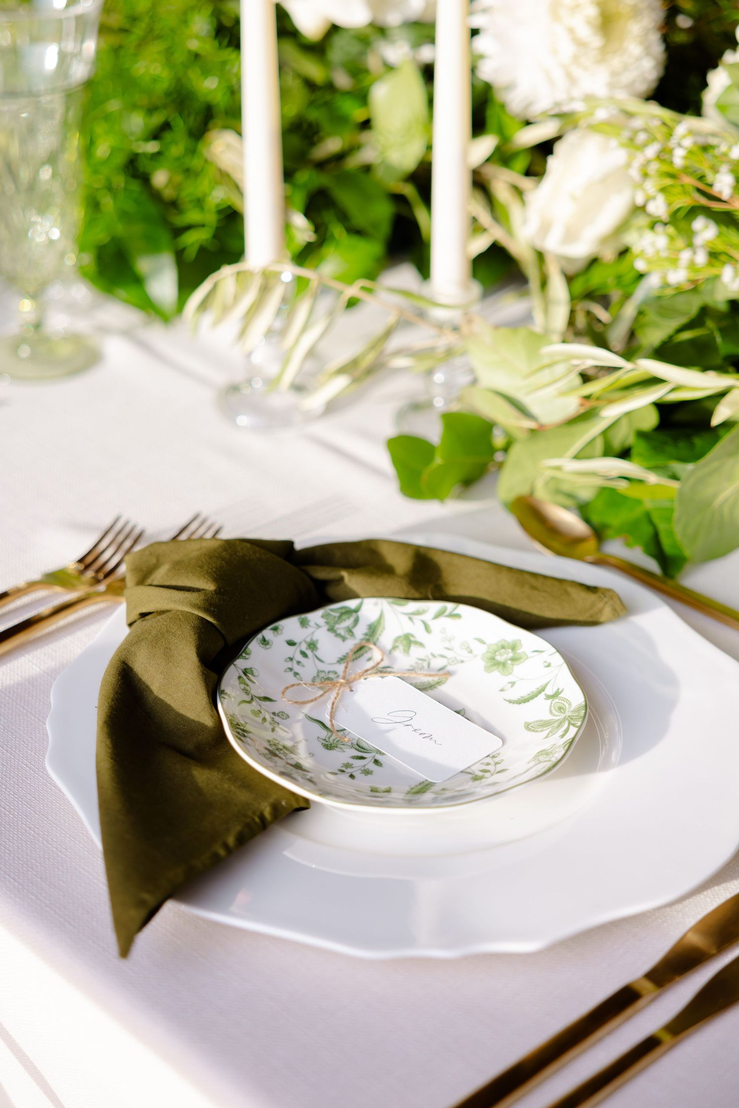 Formal place setting with white plate, olive napkin, and floral patterned accent plate.