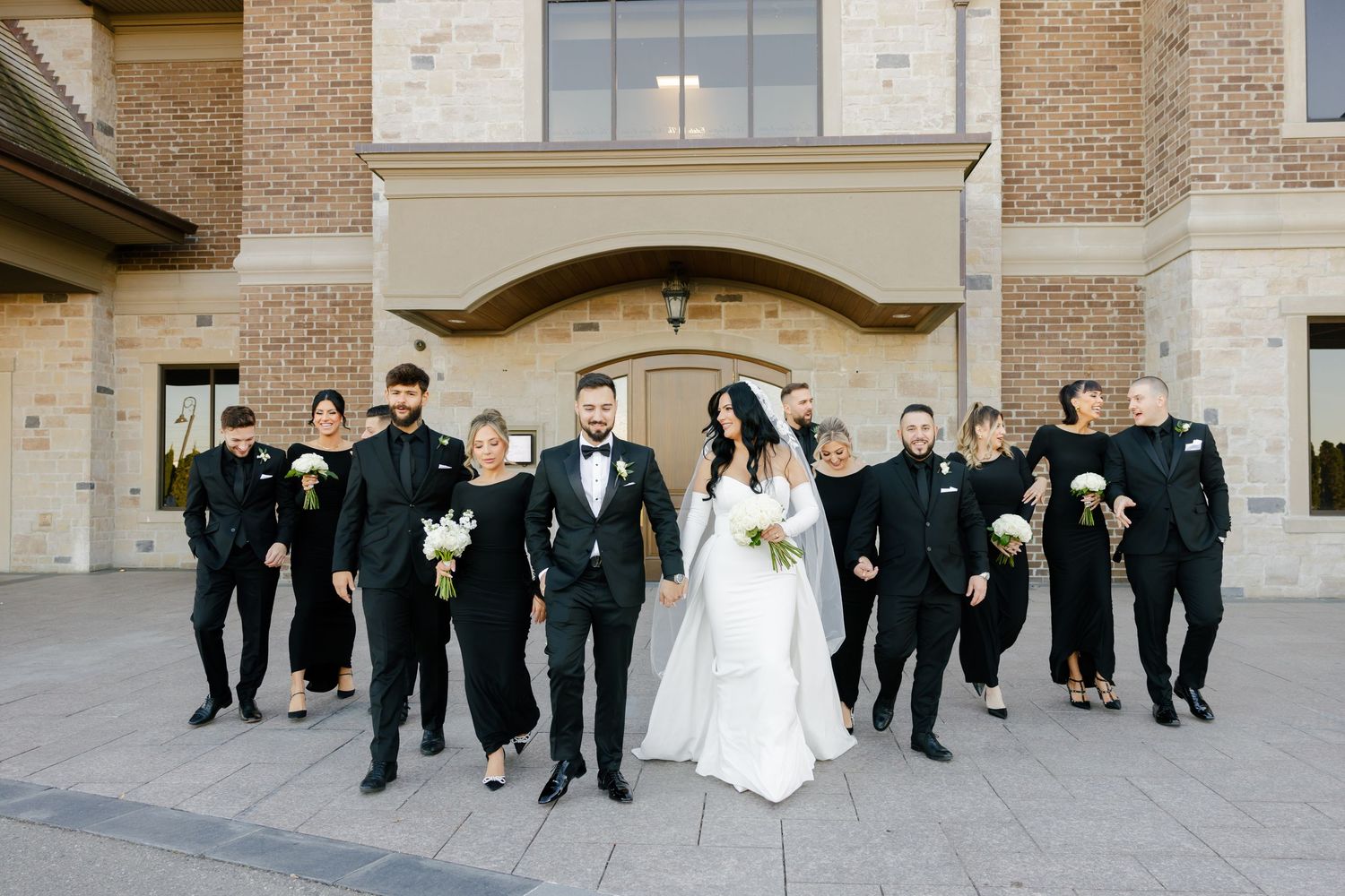 Modern wedding party walks together outside brick building with bride and groom leading the group.