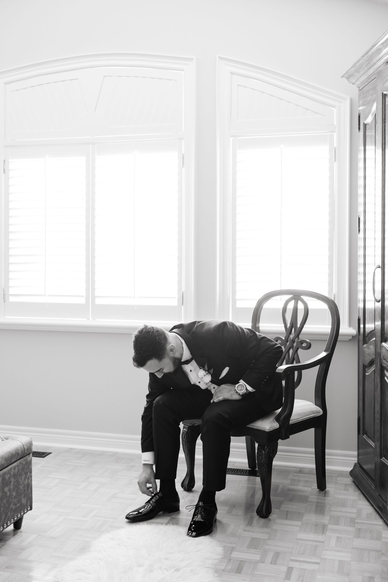 A person in formal attire sits on an antique chair while adjusting their shoe in a bright room.