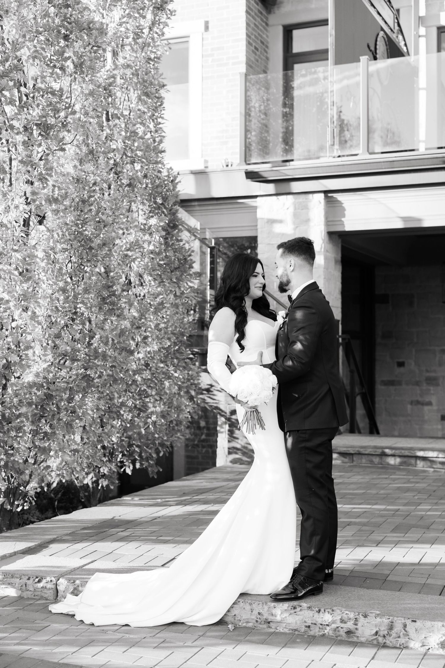 Black and white wedding portrait of couple embracing in front of modern building with autumn tree.