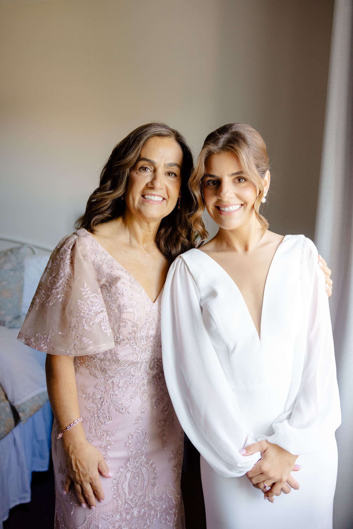 Two people smile warmly while posing together in formal dresses near a window.