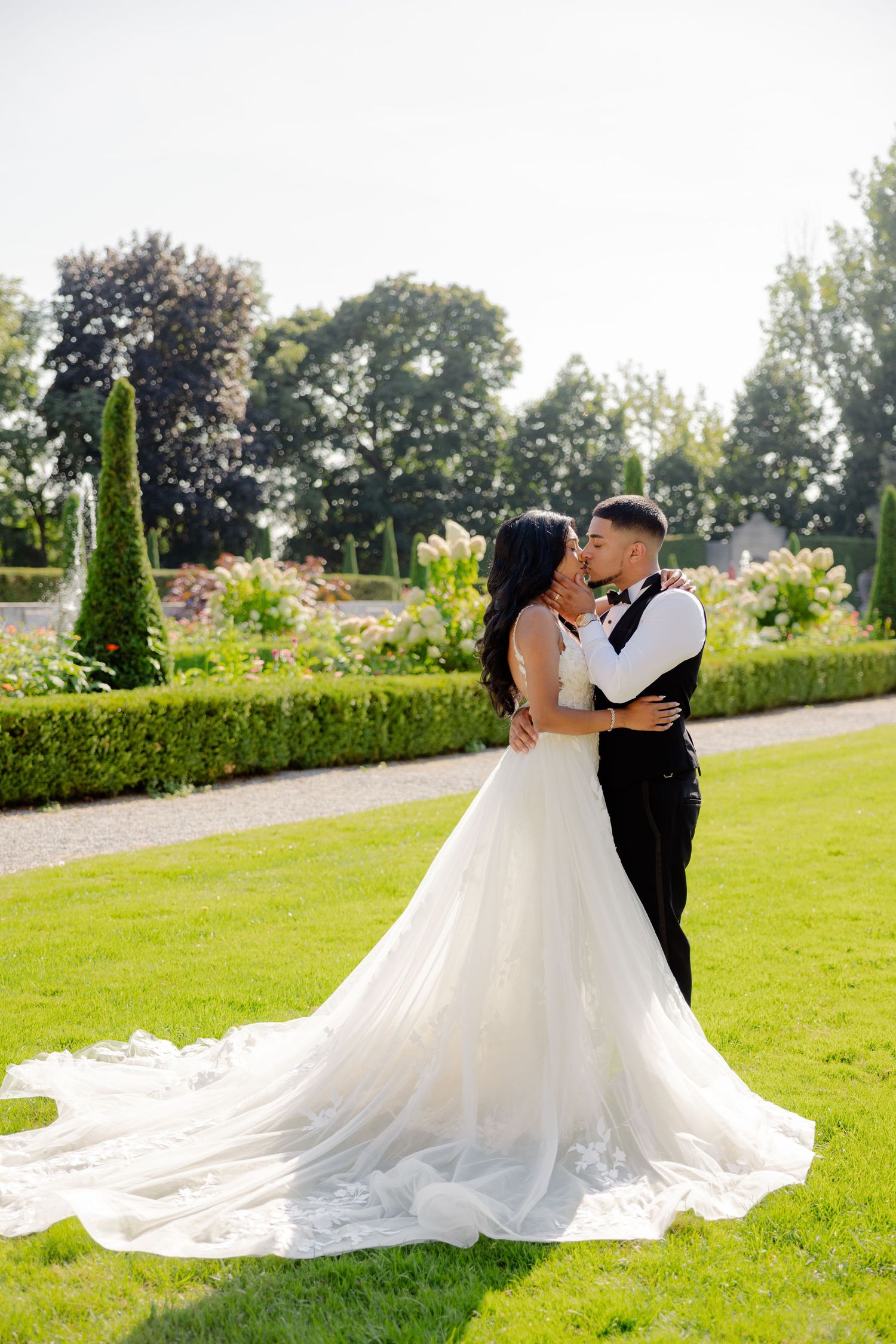 Wedding couple embraces on a pristine lawn surrounded by formal garden landscaping.