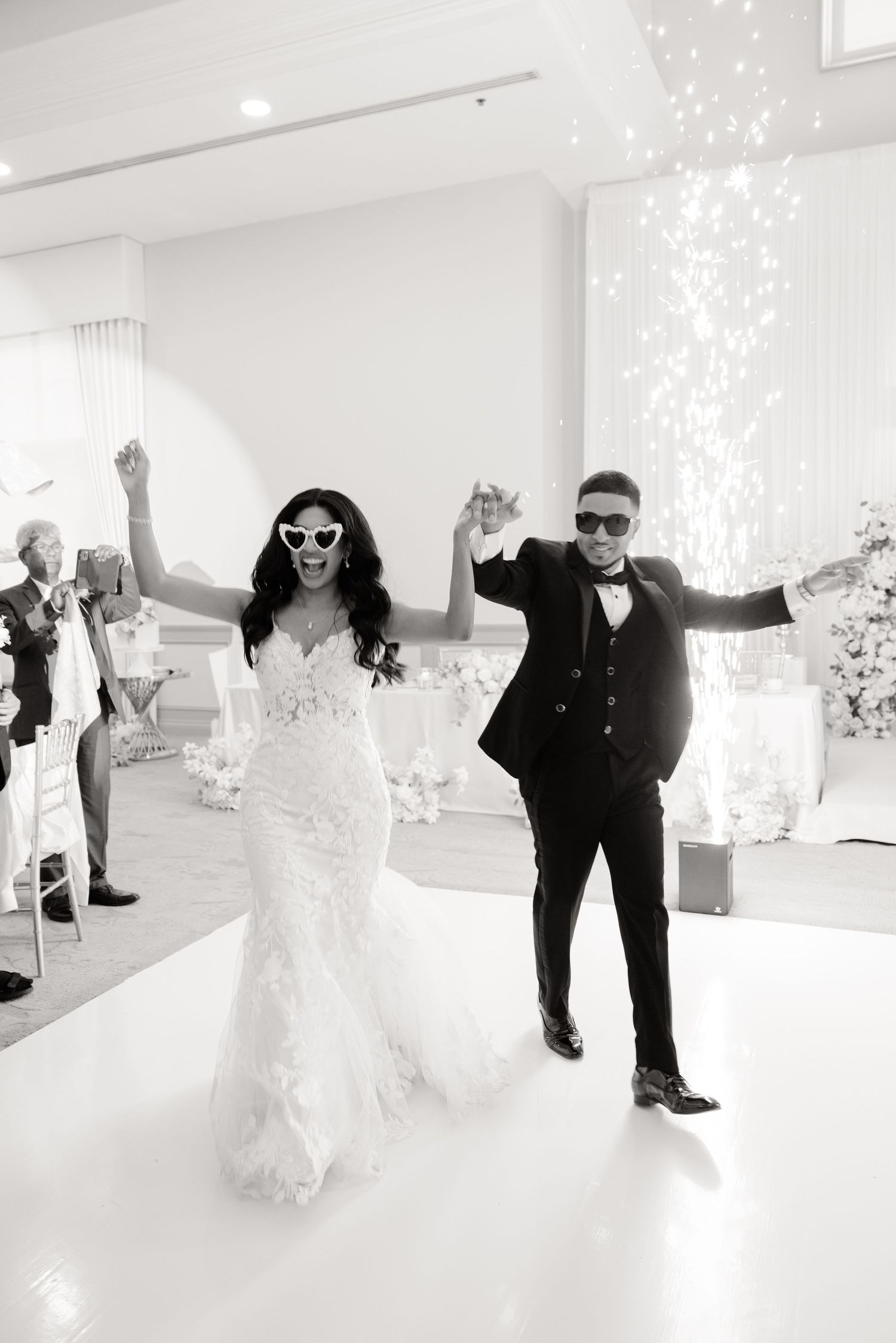 Dancing newlyweds in sunglasses celebrate on a white dance floor with sparkling lights in the background.