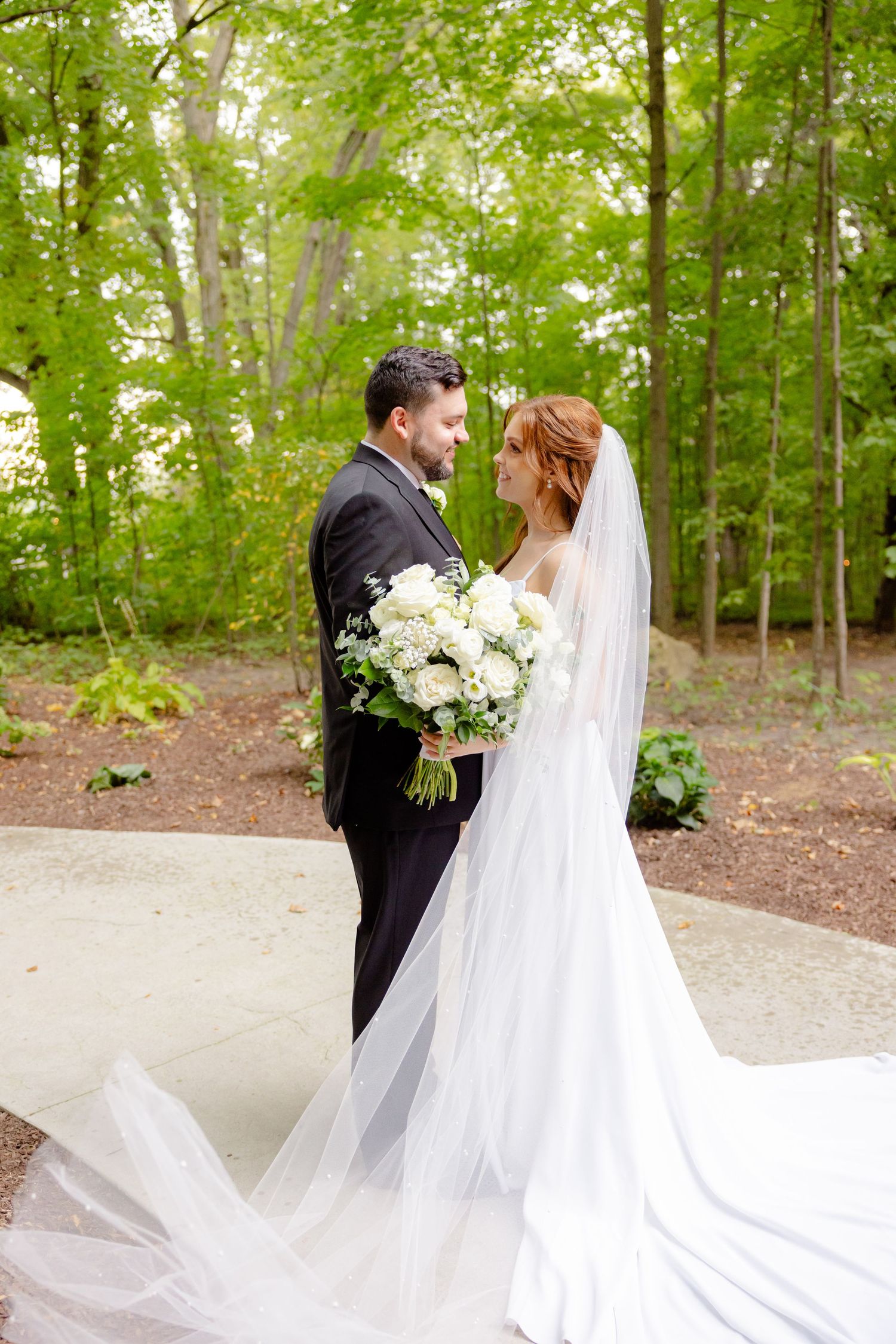 Wedding couple poses with white floral bouquet against lush green forest backdrop.
