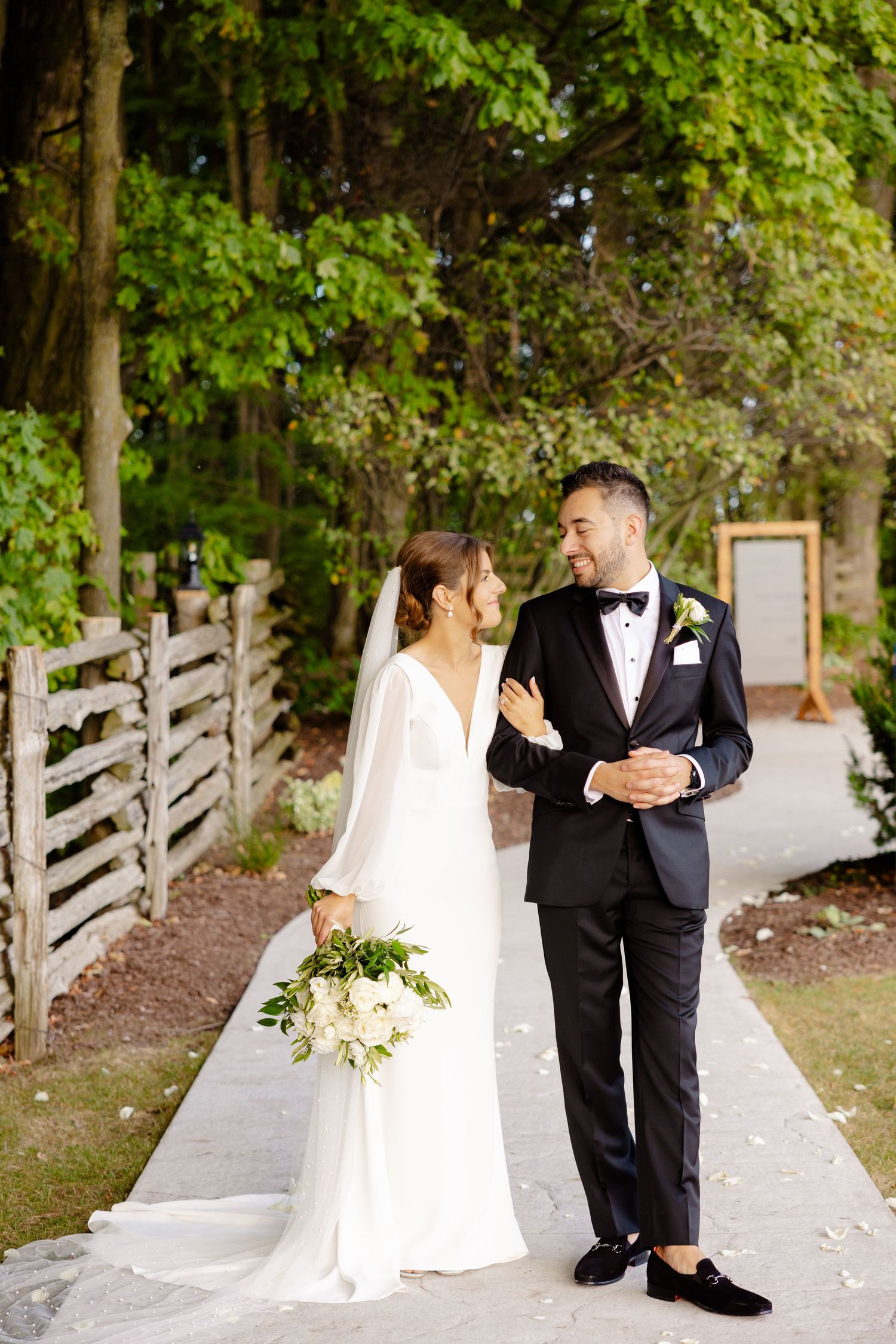 A newlywed couple shares a moment on a concrete path with rustic wood fencing and greenery.