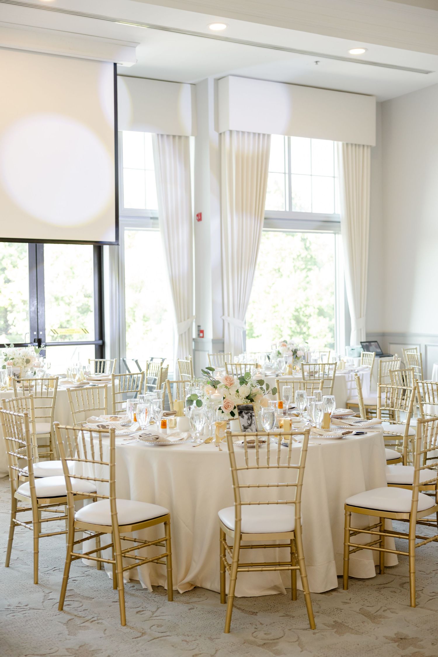Light-filled wedding reception venue with round tables, gold chiavari chairs, and ivory linens.