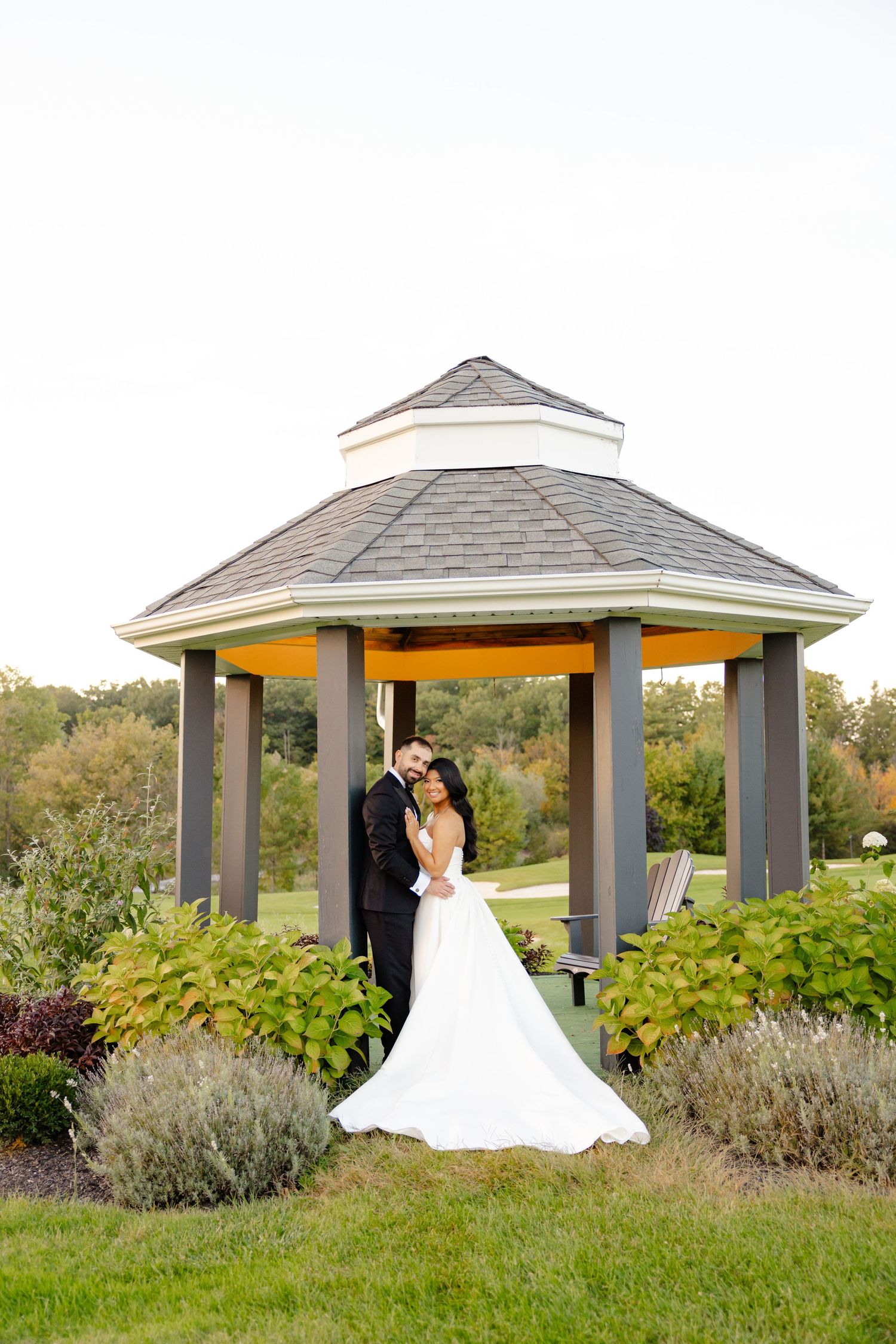 A romantic wedding portrait of a couple standing in an elegant outdoor gazebo with landscaped gardens.