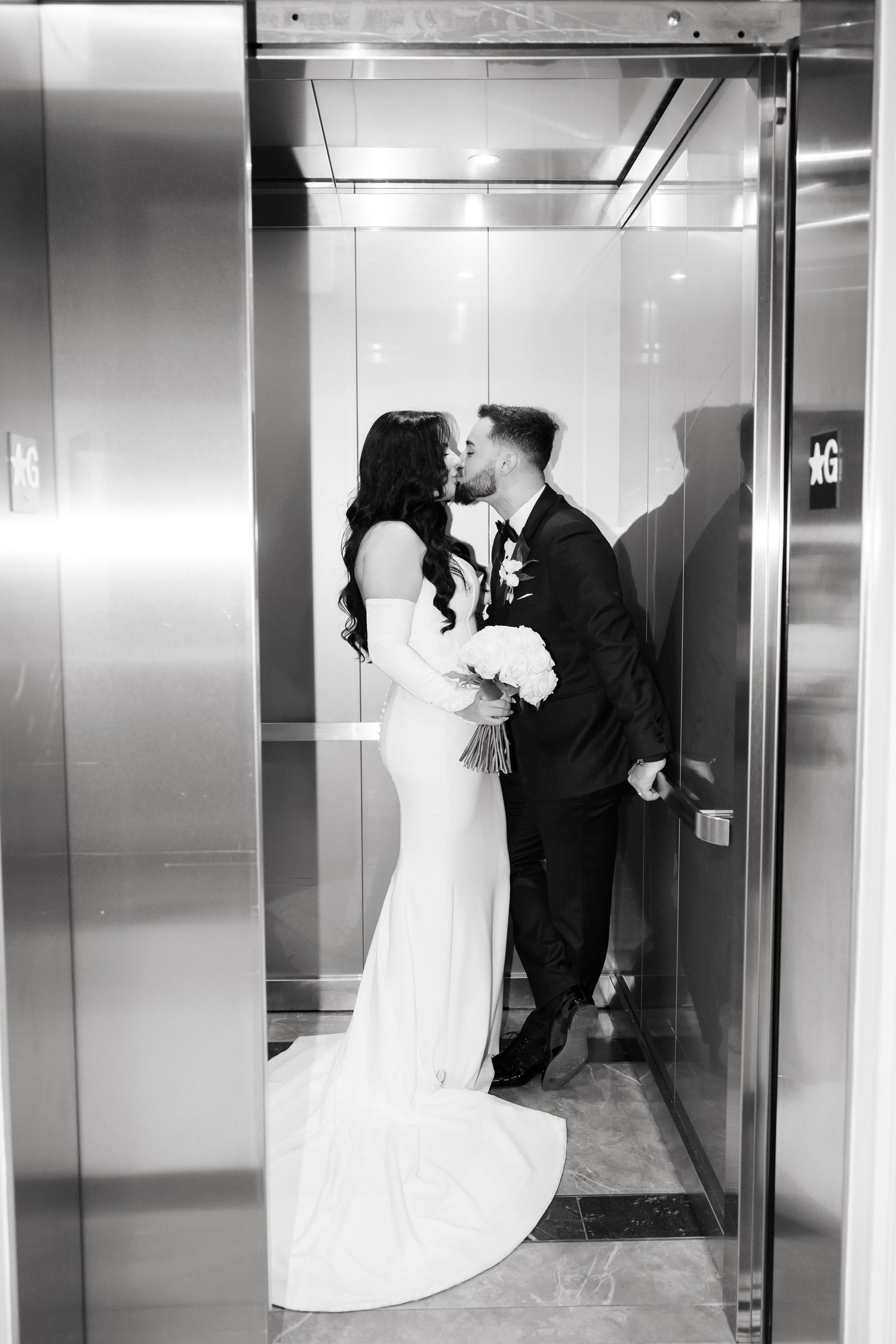 Black and white wedding photo capturing intimate kiss in modern elevator setting.