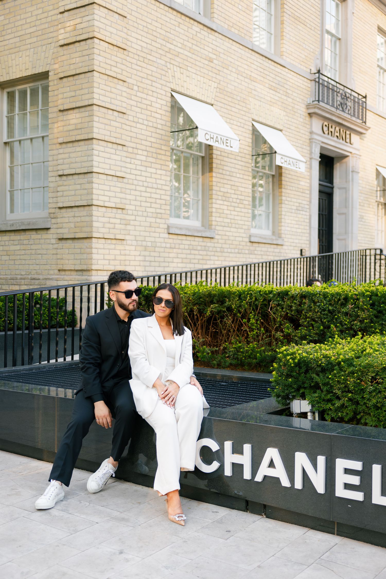 Fashion-forward pair in monochrome outfits sitting on Chanel store signage.