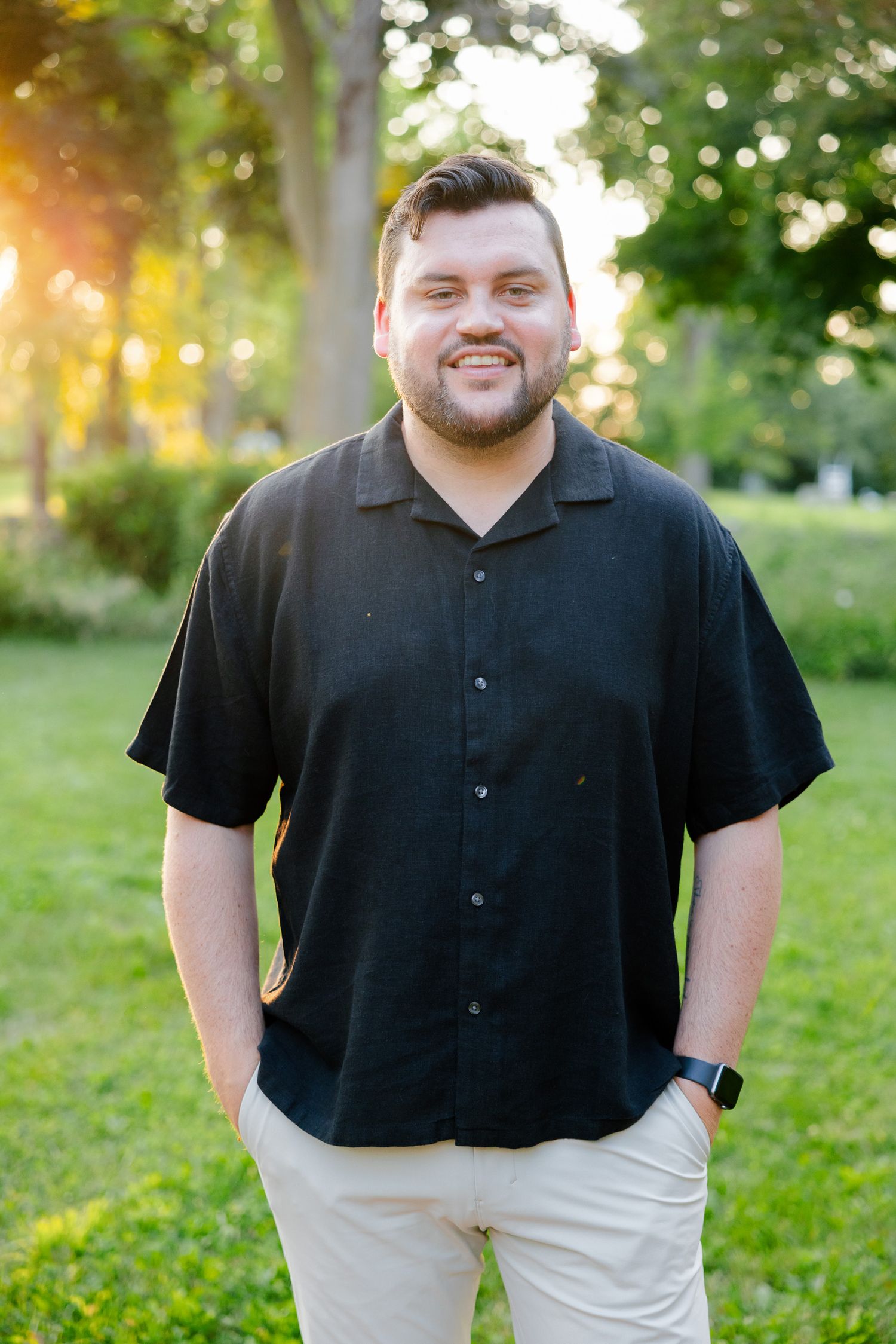 Portrait in a park setting with warm sunset lighting through trees wearing a black button-up shirt.