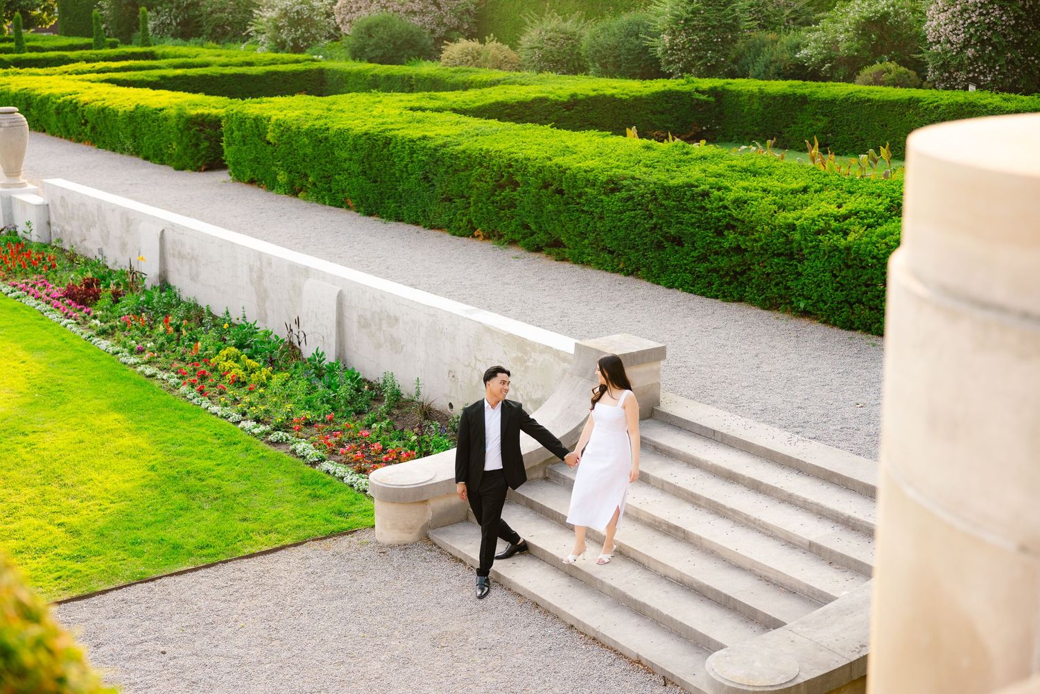 Two people sit casually on curved stone steps in a formal garden with meticulously trimmed hedges.