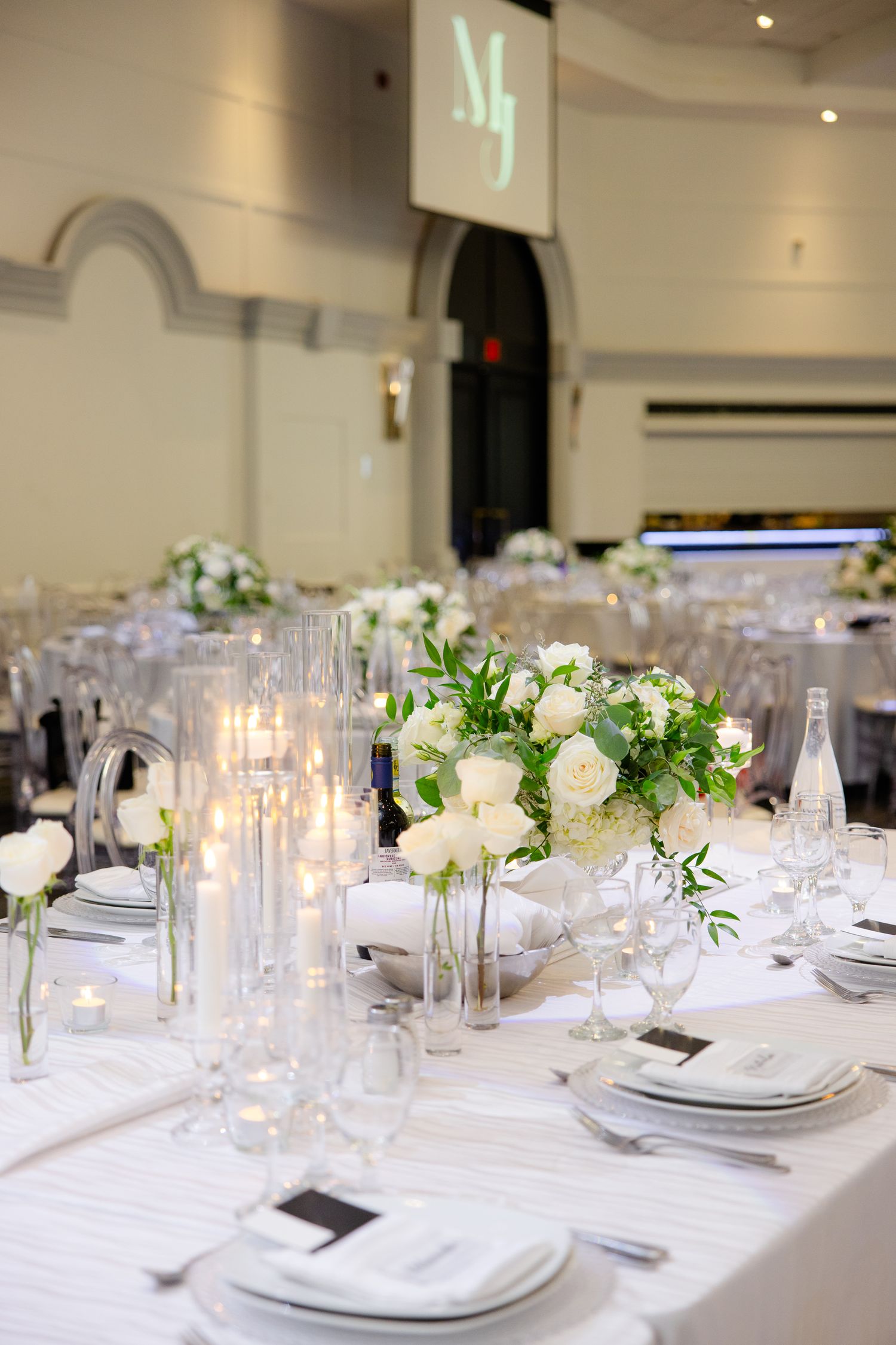 Modern wedding reception tables with white floral centerpieces and candles in an elegant ballroom setting.