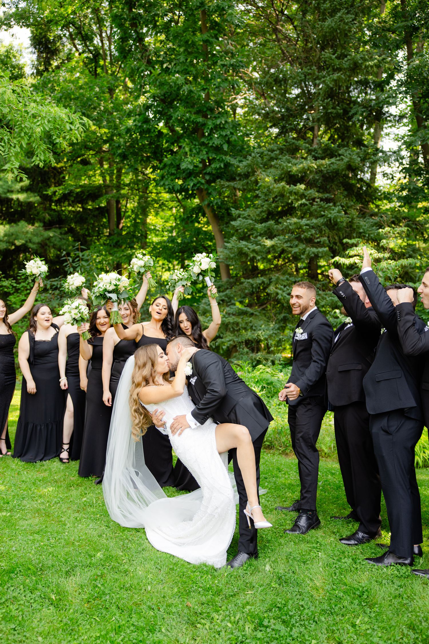 Wedding party cheers as couple shares dramatic dip kiss on grass with forest background.