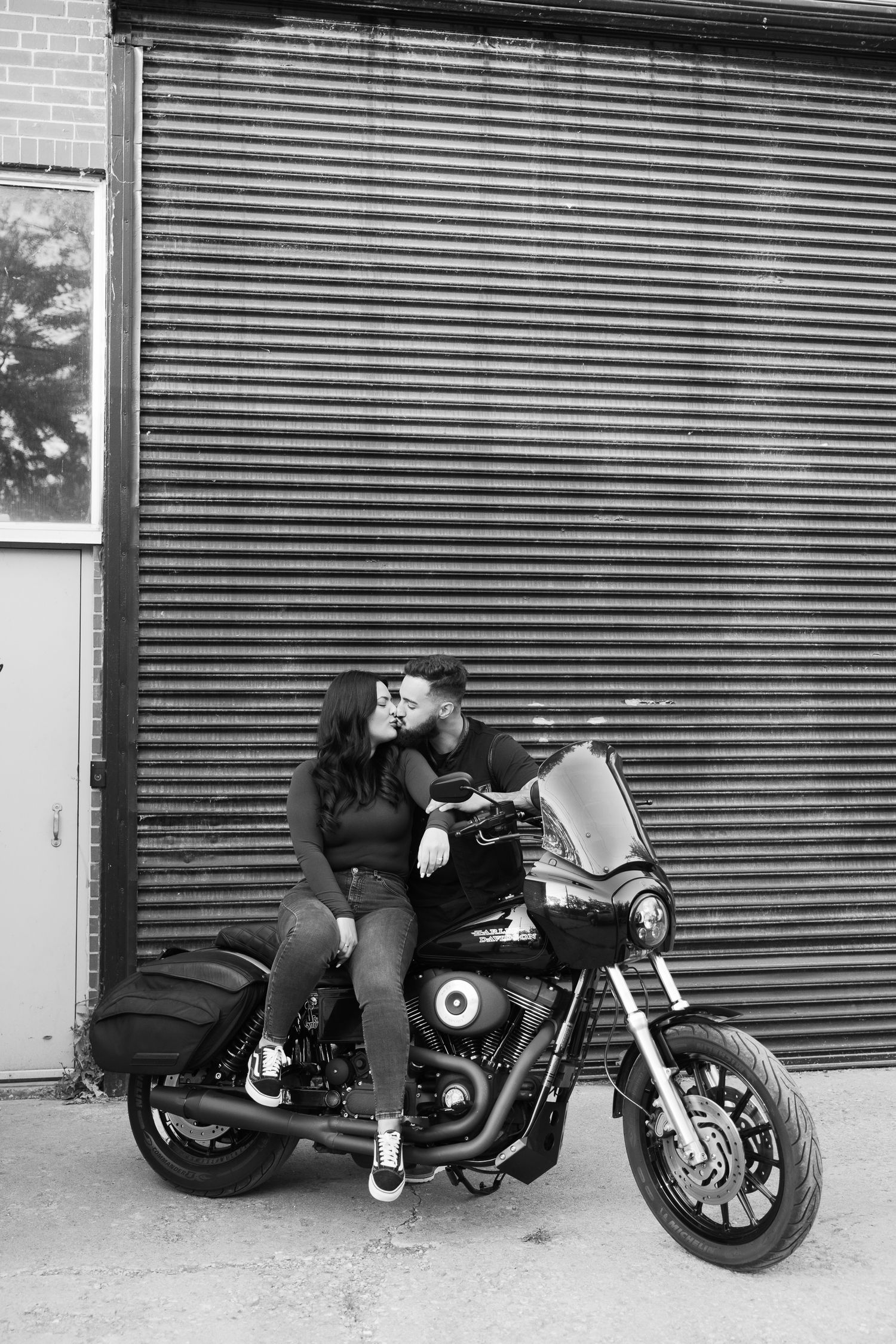 A couple sits intimately on a black motorcycle against a corrugated metal wall in black and white.