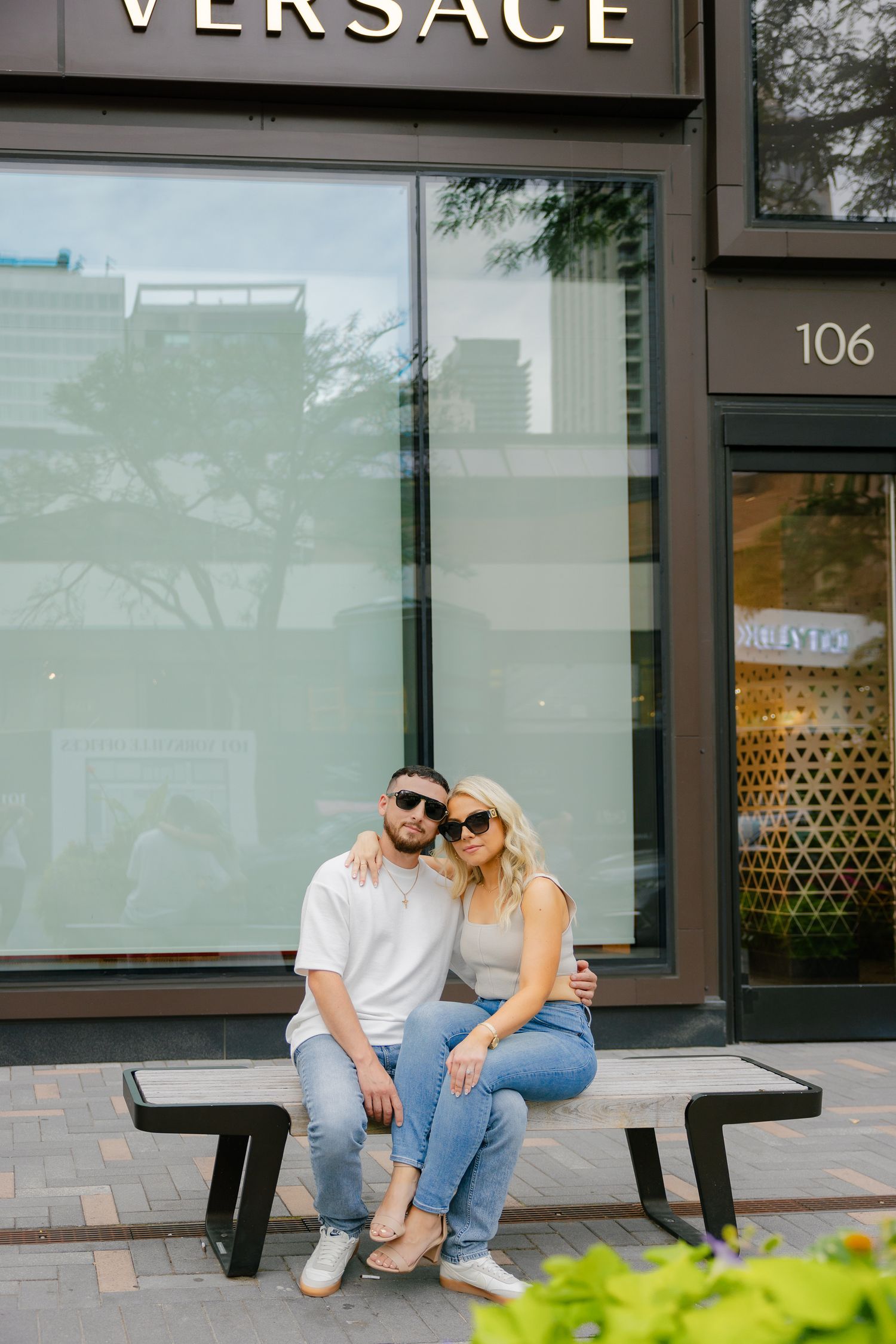 Couple relaxing on bench outside Versace store wearing coordinated casual outfits.