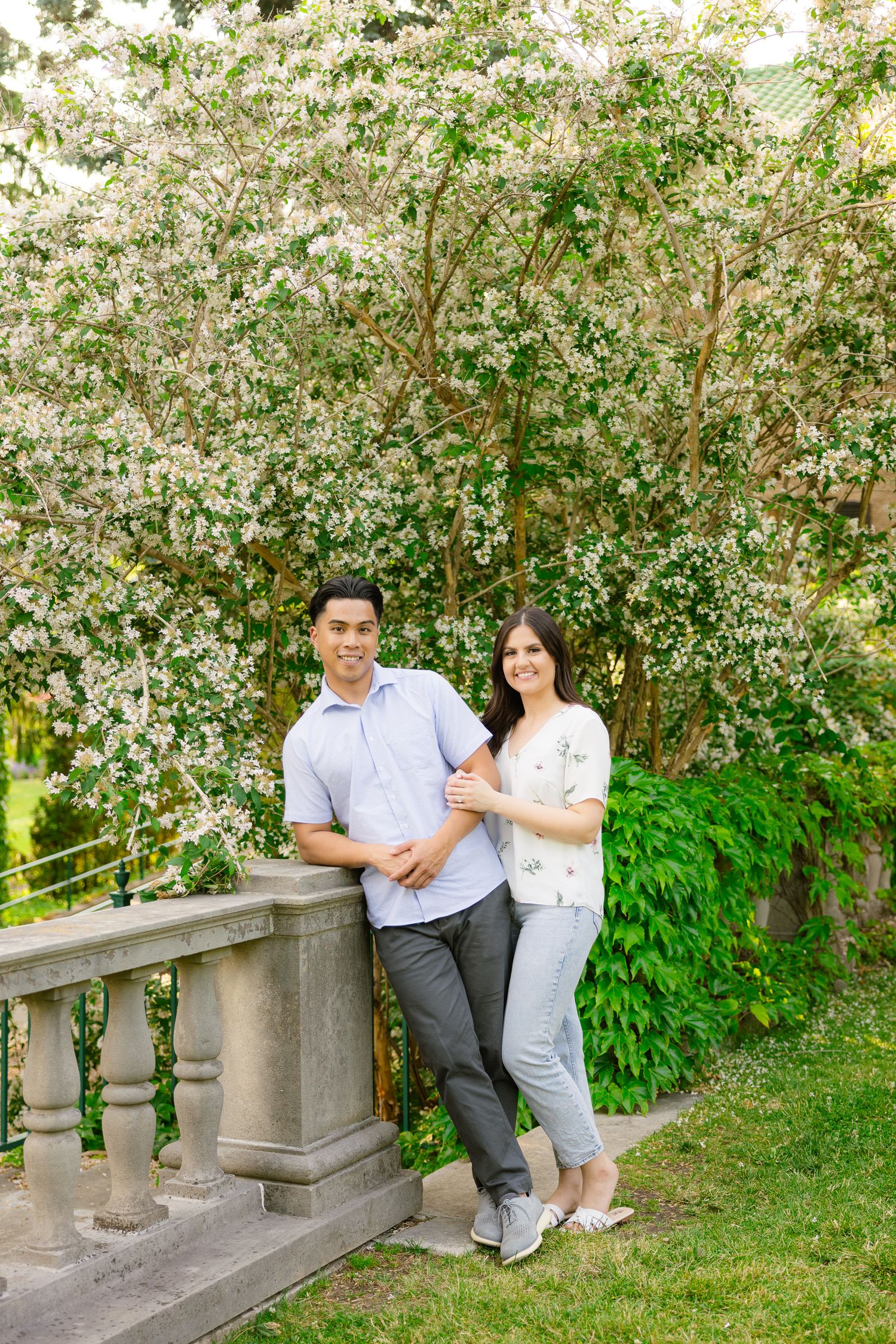 Couple poses by stone railing with blooming white flowers and greenery in background.