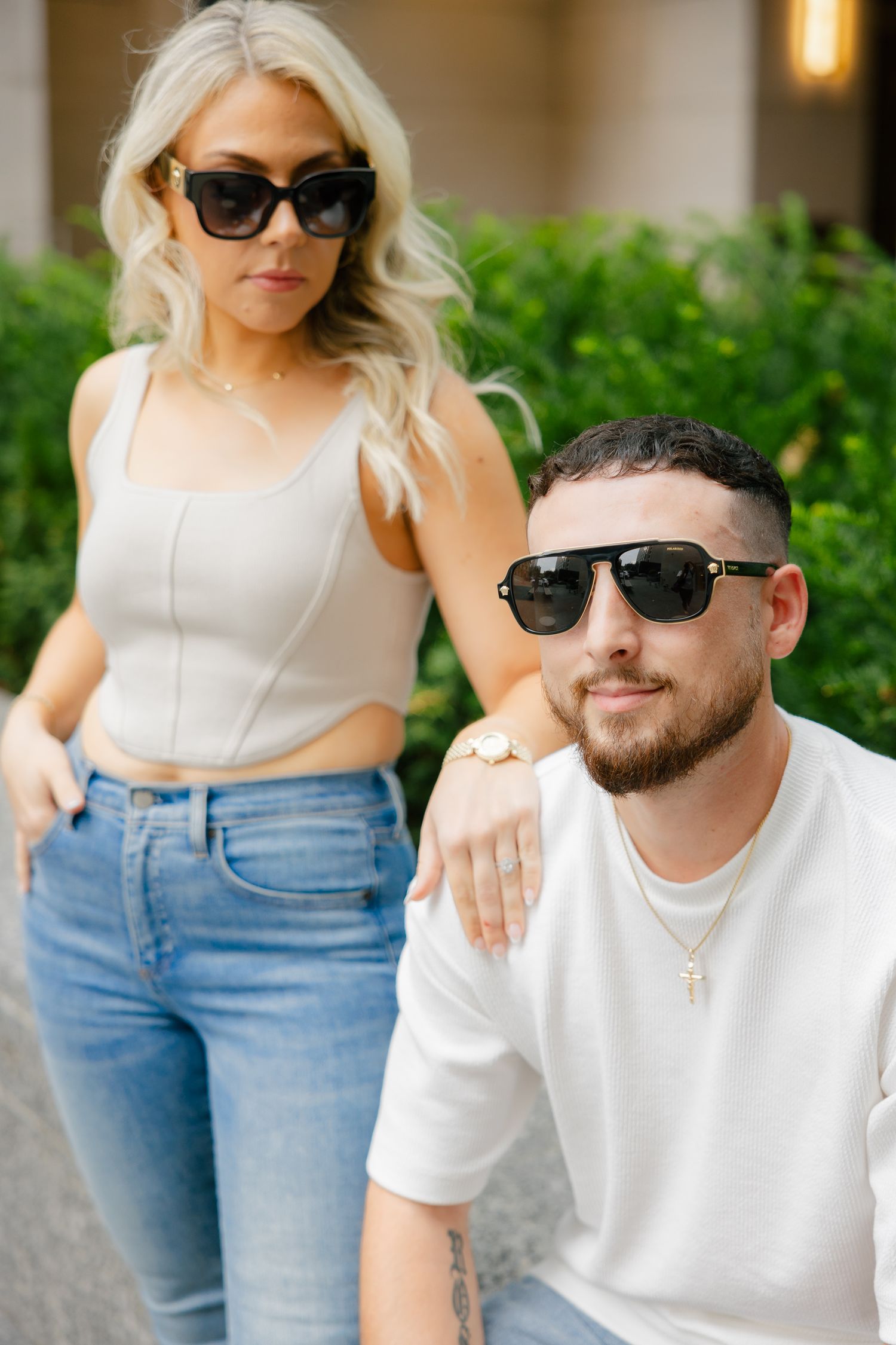 Fashion-forward couple wearing sunglasses pose against urban greenery backdrop.