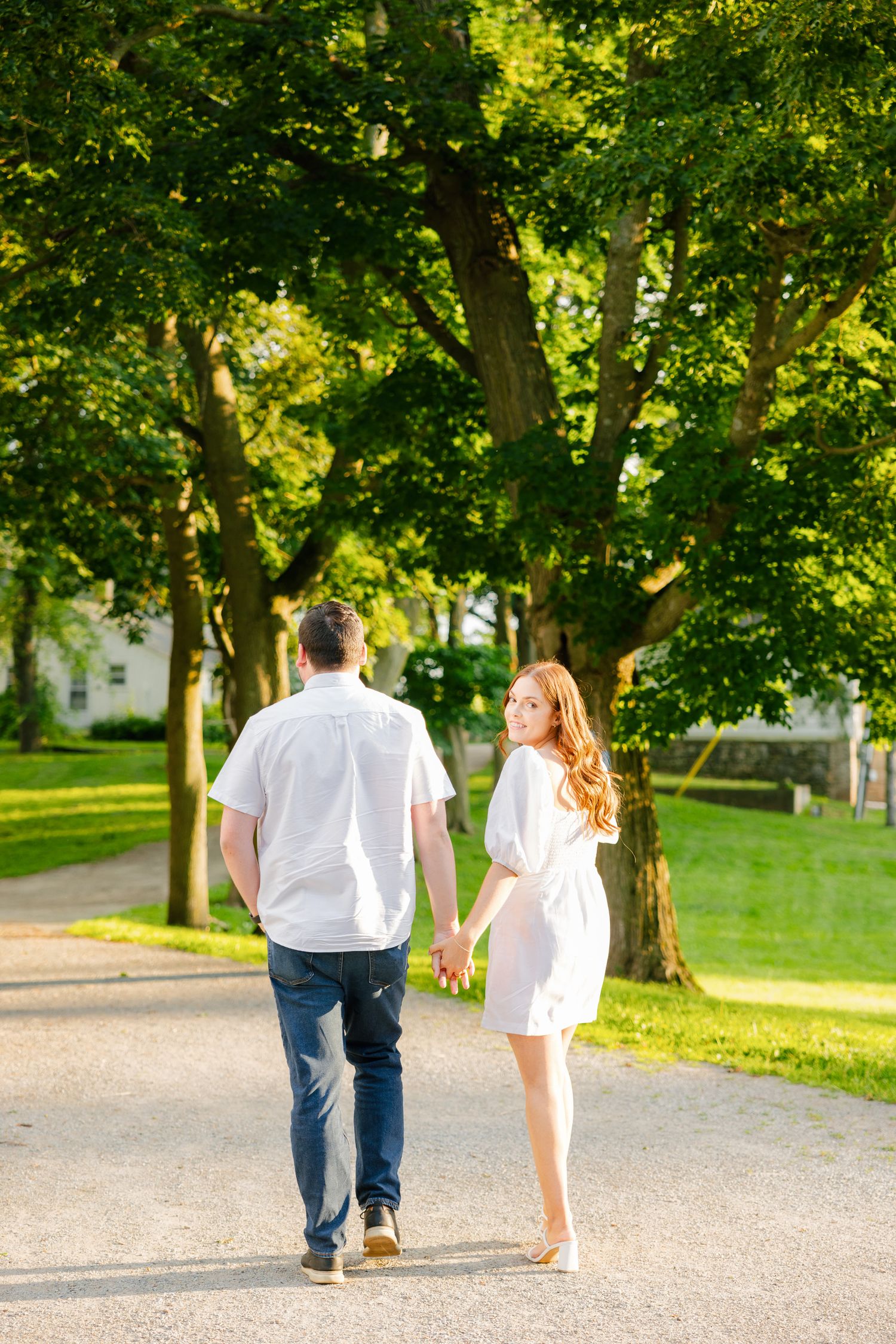 A couple in white and denim walking hand in hand along a tree-lined path on a sunny day.