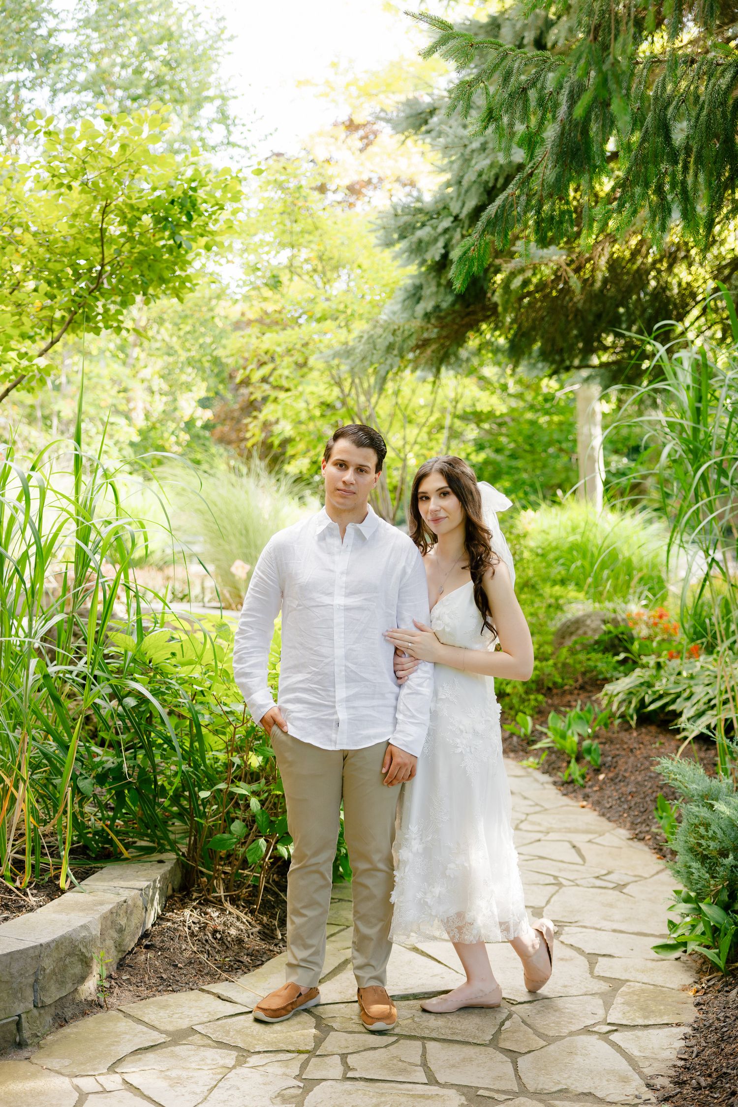 A couple stands together on a stone pathway in a vibrant garden setting.