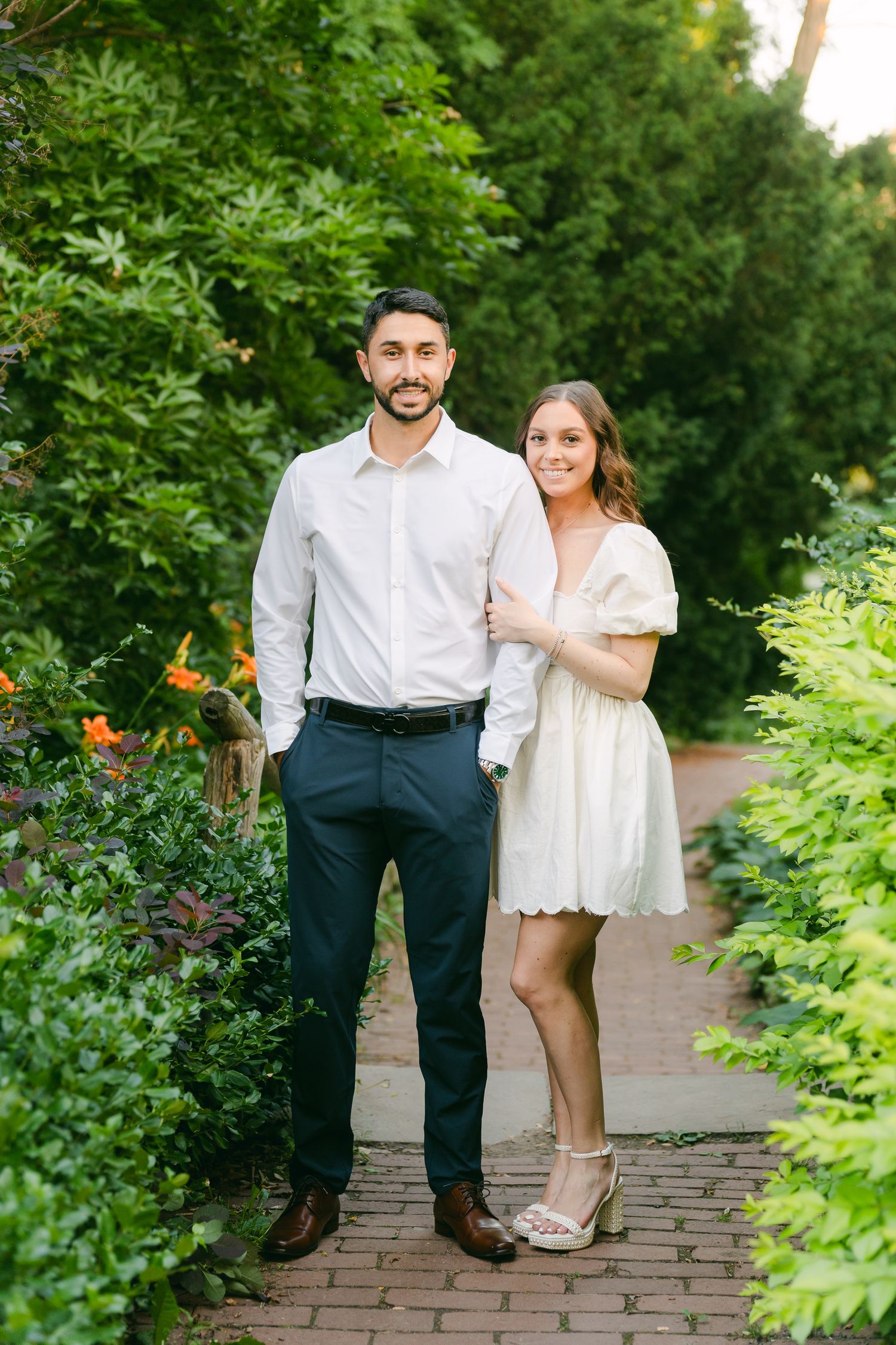 Elegantly dressed couple posing on brick pathway surrounded by colorful garden flowers.