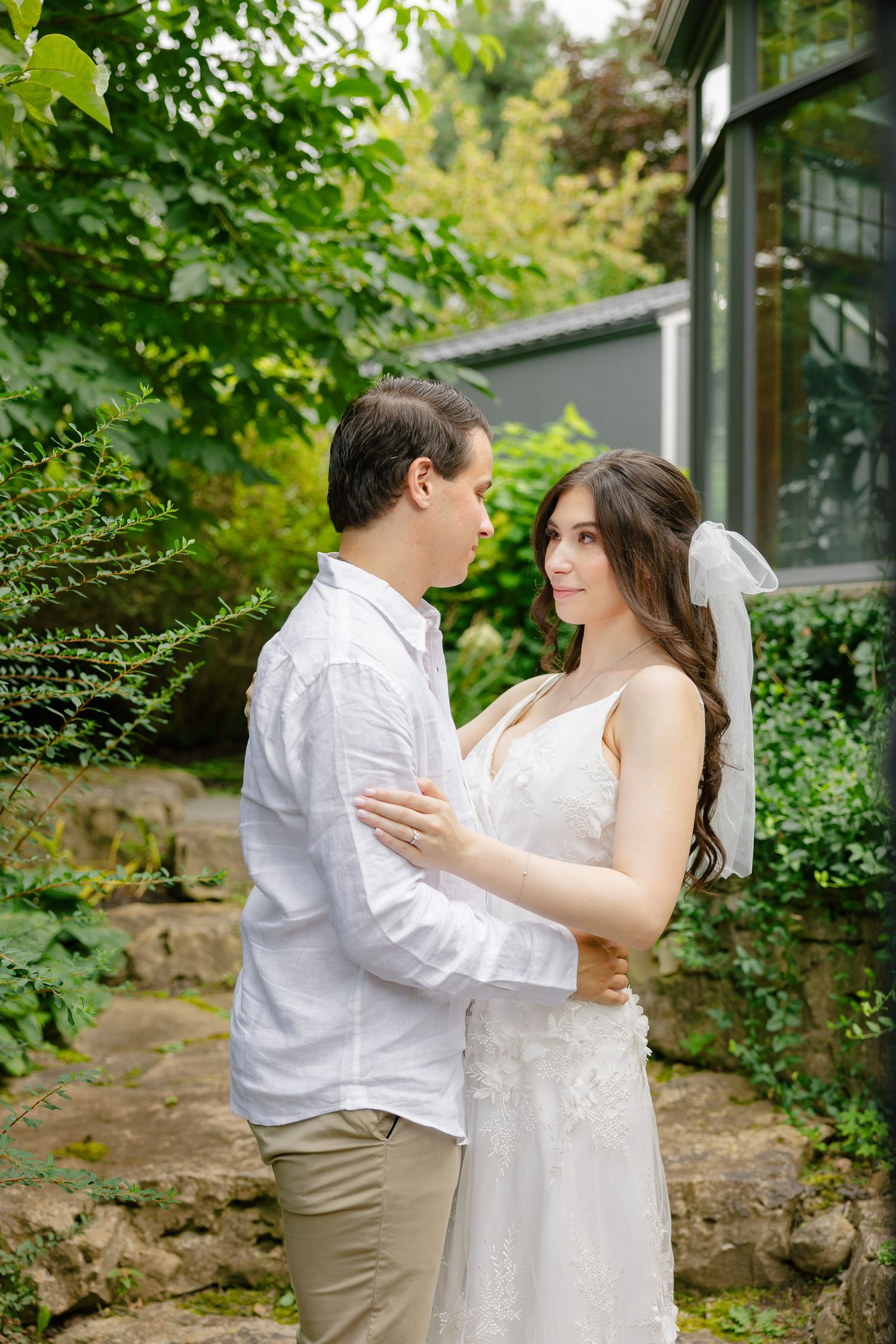 Wedding couple in casual elegance poses against a greenhouse backdrop surrounded by greenery.
