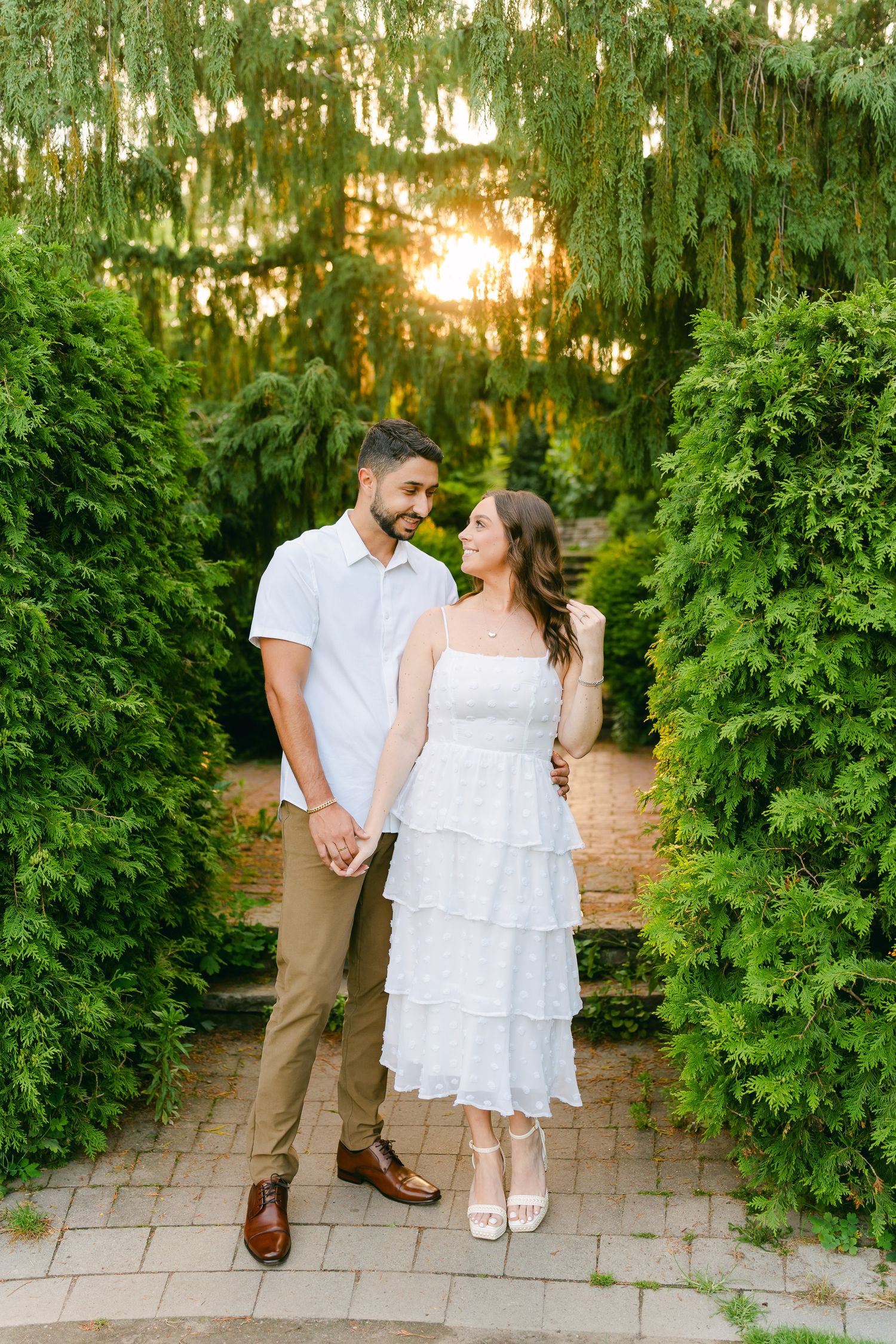 Couple walks hand in hand down brick steps between manicured hedges at sunset.