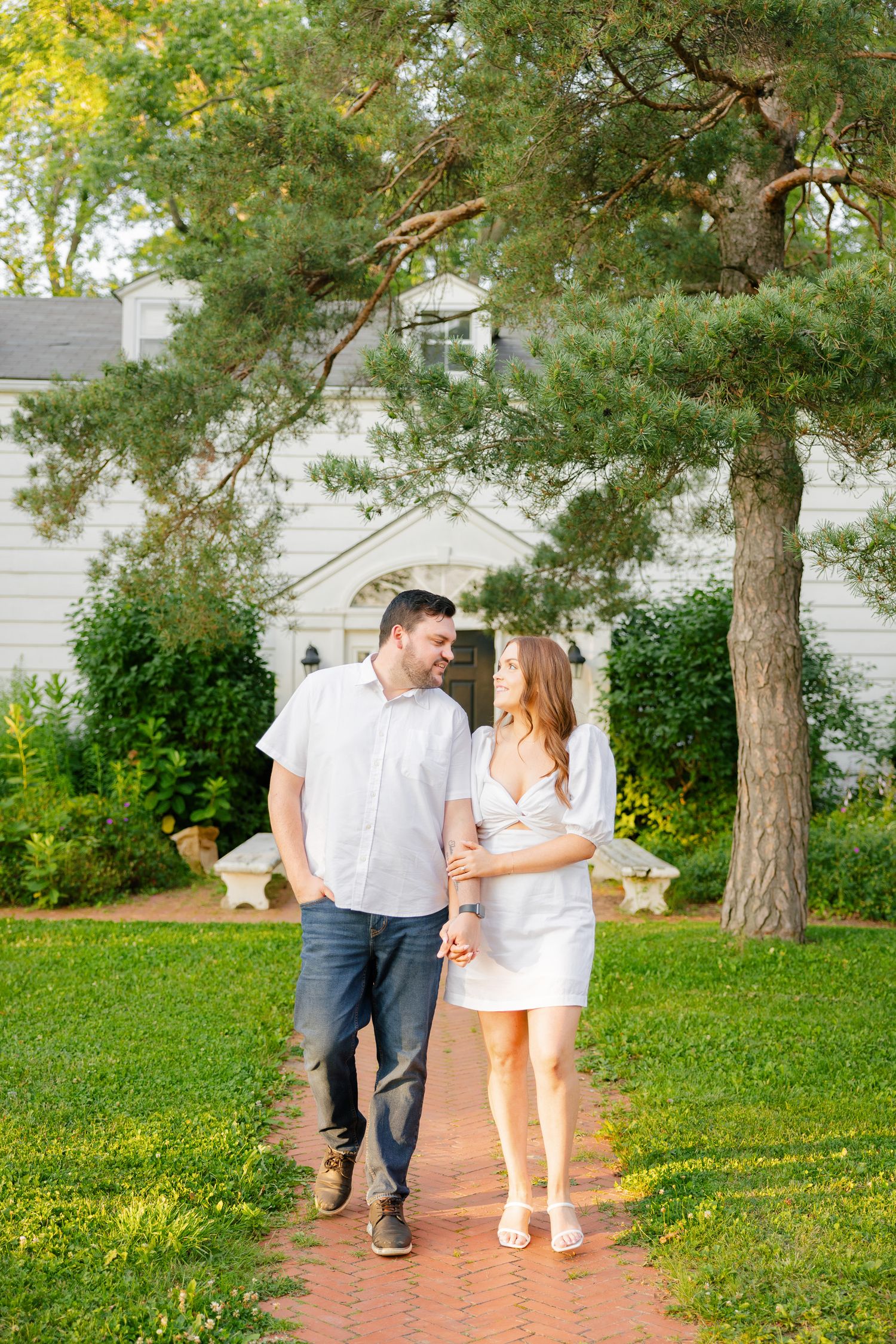 A couple strolls together on a brick pathway surrounded by green lawn and pine trees.