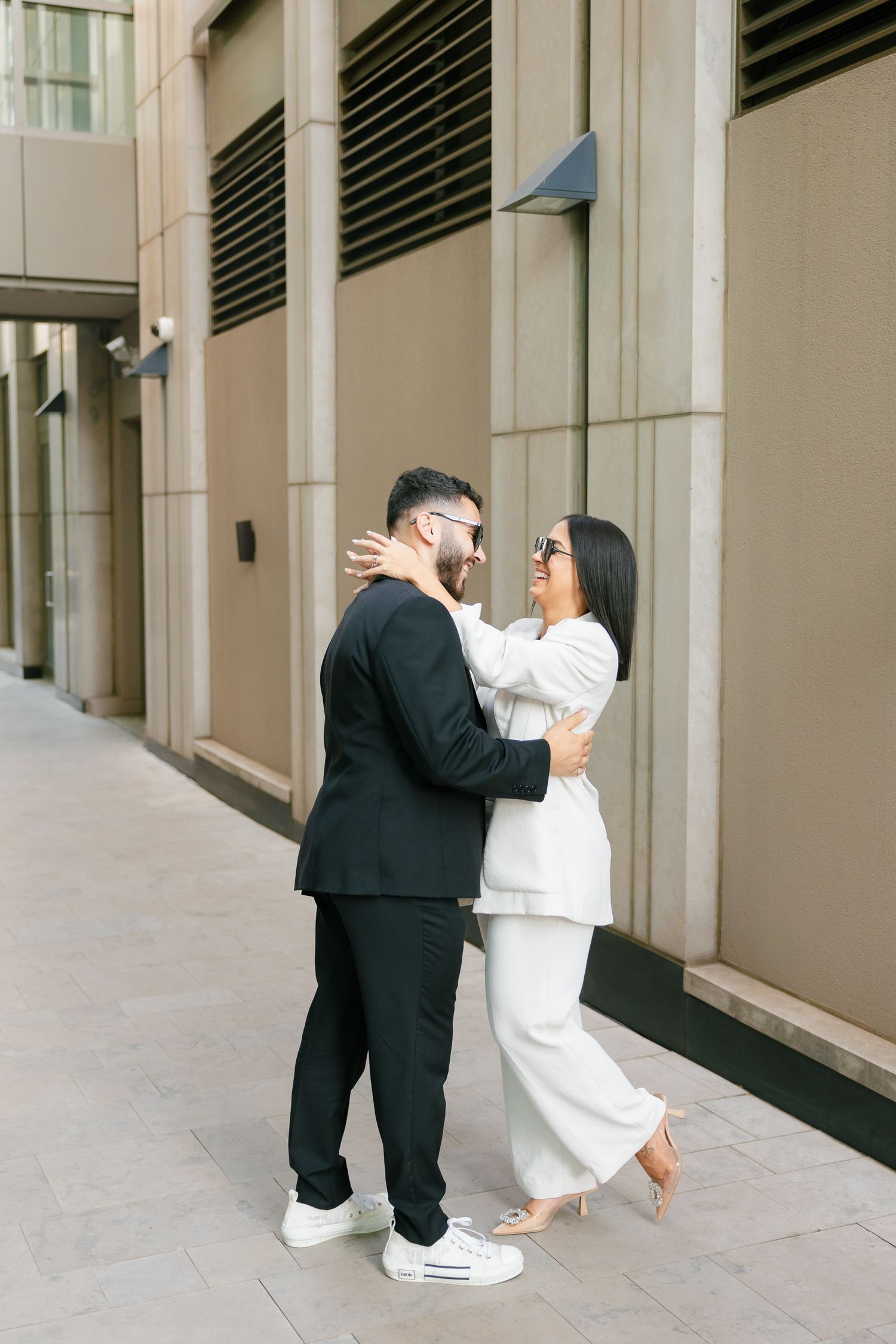 A couple in contrasting black and white outfits embraces on a city sidewalk outside a modern building.